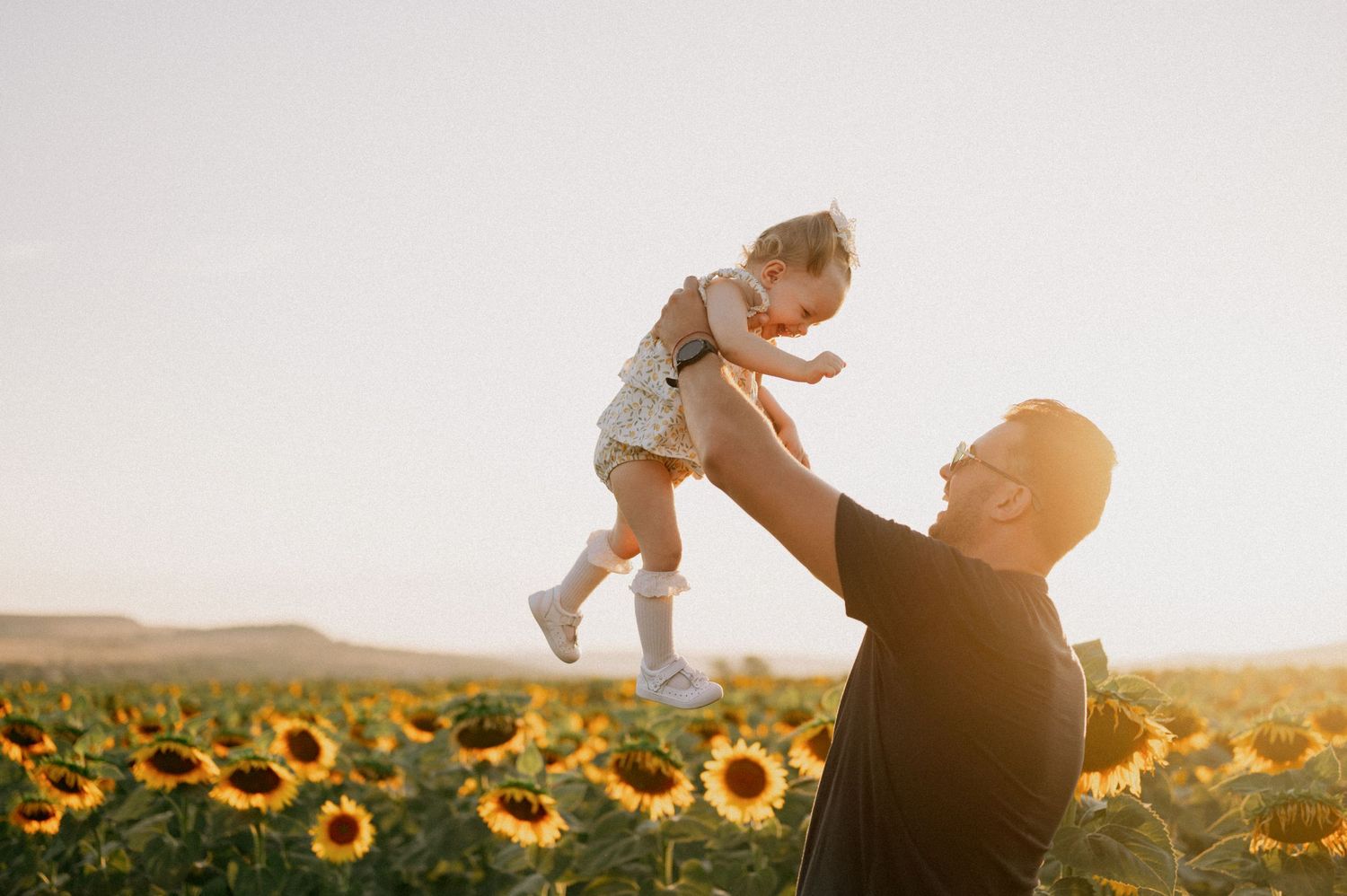 Man lifting toddler girl above sunflower field in golden sunset light, joyful father-daughter moment outdoors.
