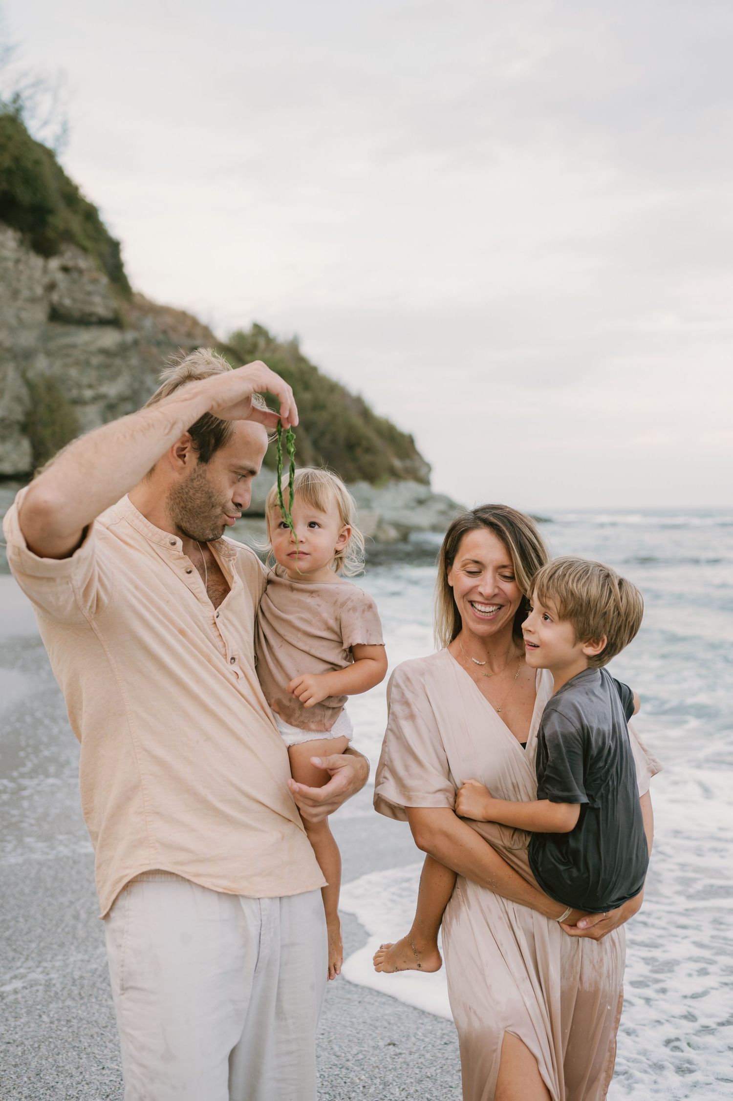Family of four enjoying a beach day, parents holding young children near ocean waves with rocky cliffs in background.