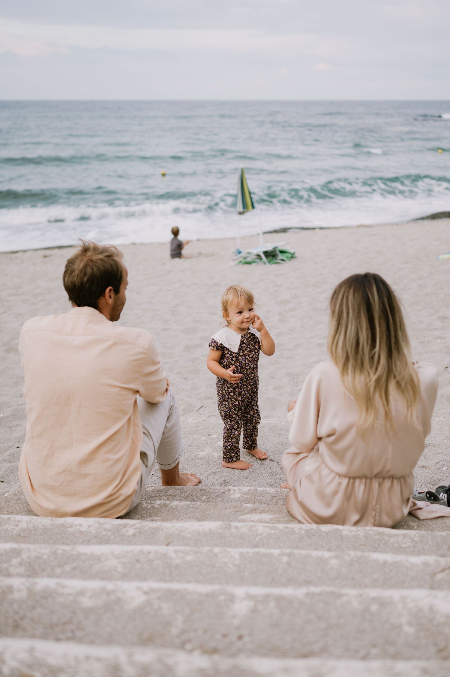 Family sitting on beach steps watching their toddler play, with ocean waves in the background.