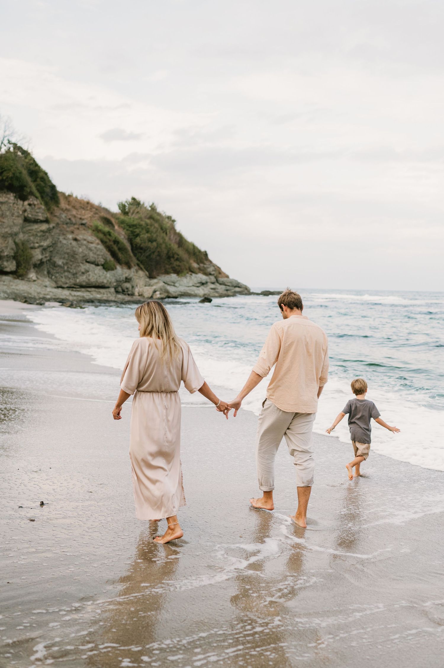 Family of three walking barefoot on a sandy beach, holding hands near ocean waves with rocky cliffs in background.