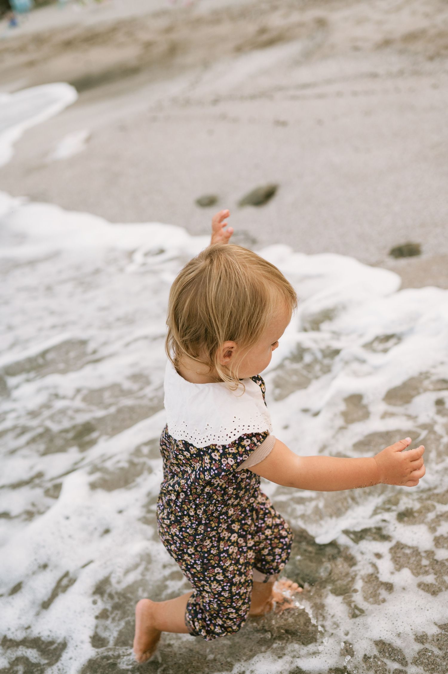 Toddler girl in floral romper playing in ocean waves on a sandy beach.