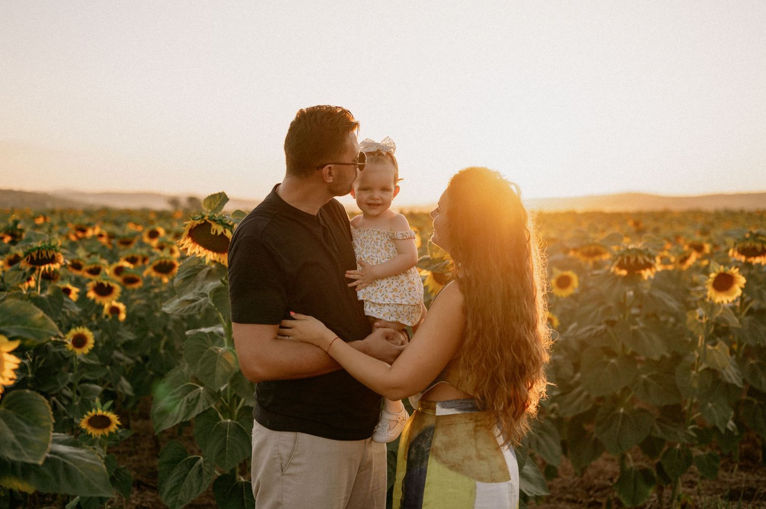 Family of three posing in a sunflower field at golden hour sunset, parents holding baby girl.