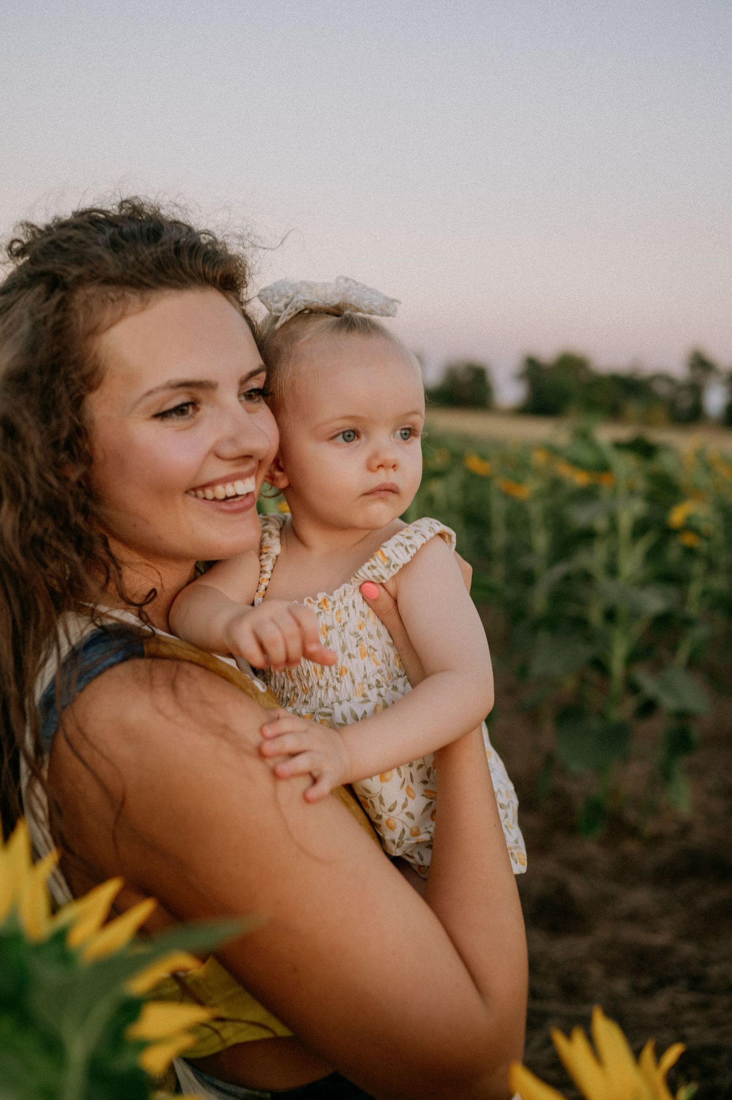 A smiling young mother holds her baby girl in a sunflower field at golden hour sunset.