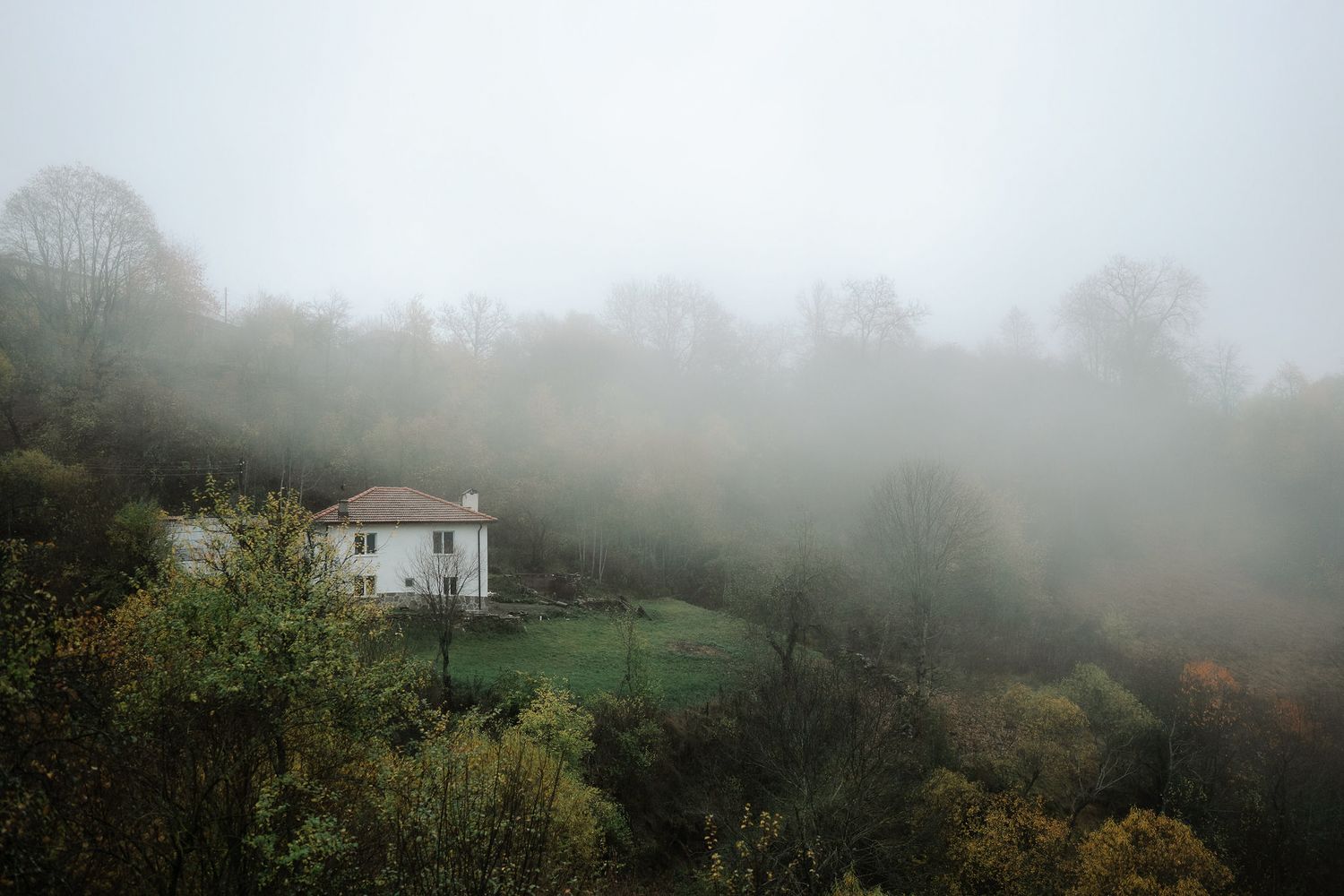 Moody landscape with fog rolling over green hills and a solitary white house in the distance.