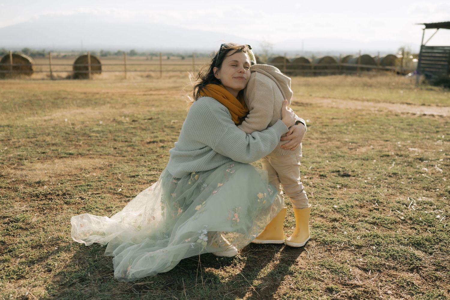 Someone in a flowy white dress kneels in a rural field while embracing a golden retriever dog.