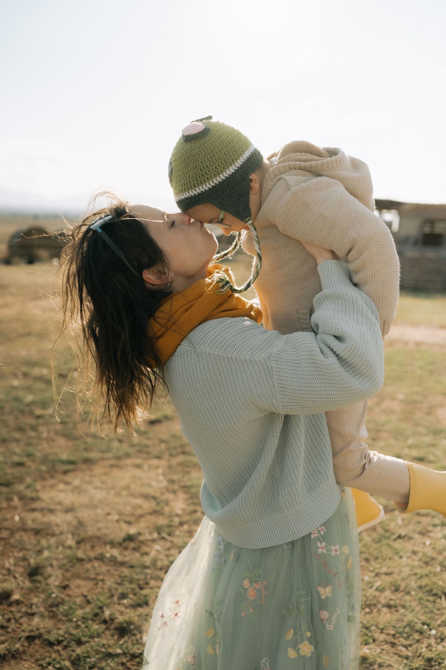 A person in a gray sweater and floral dress holds and lifts up a small dog in a sunny outdoor field.