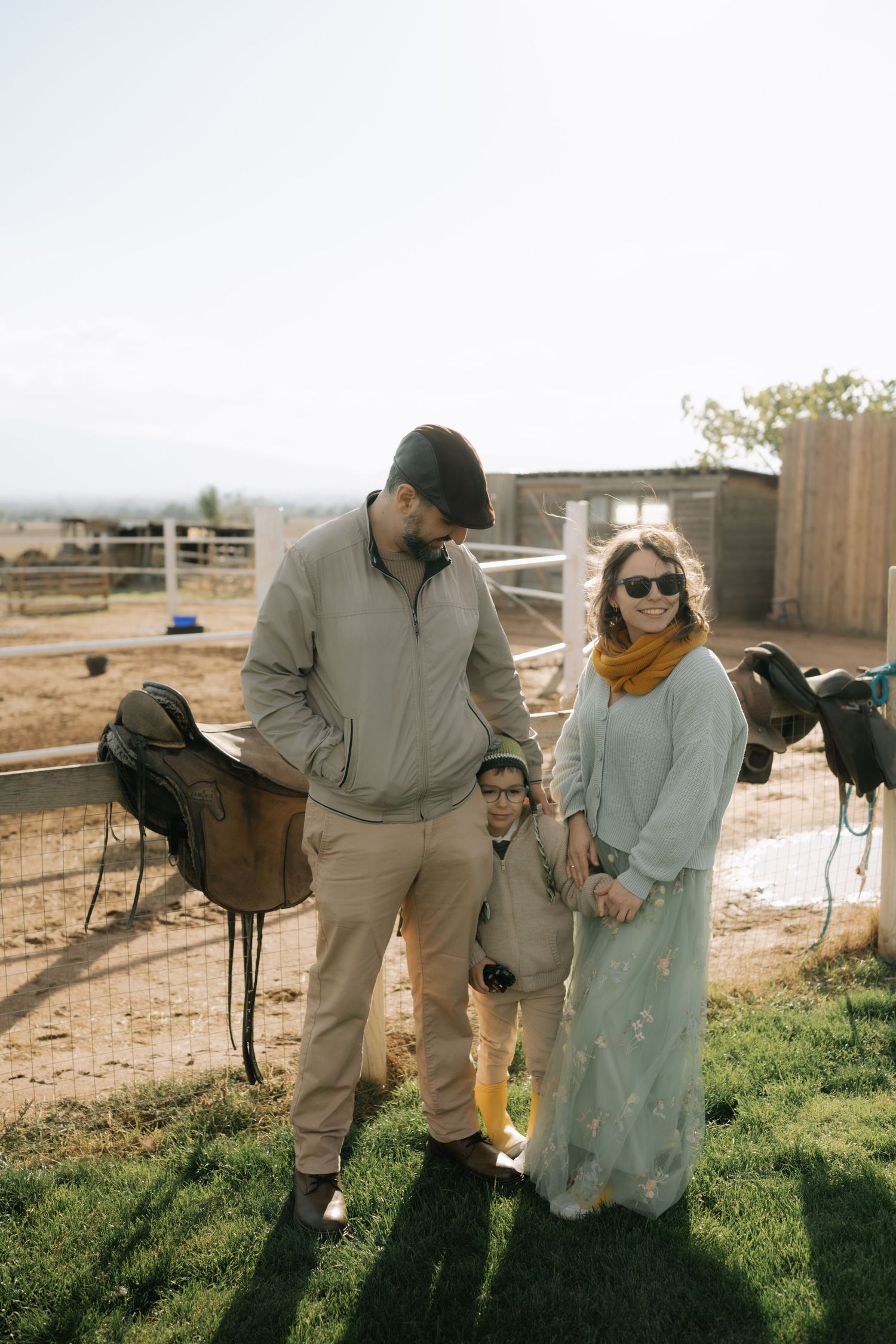 A couple walks together through a rustic outdoor setting with horses in the background.