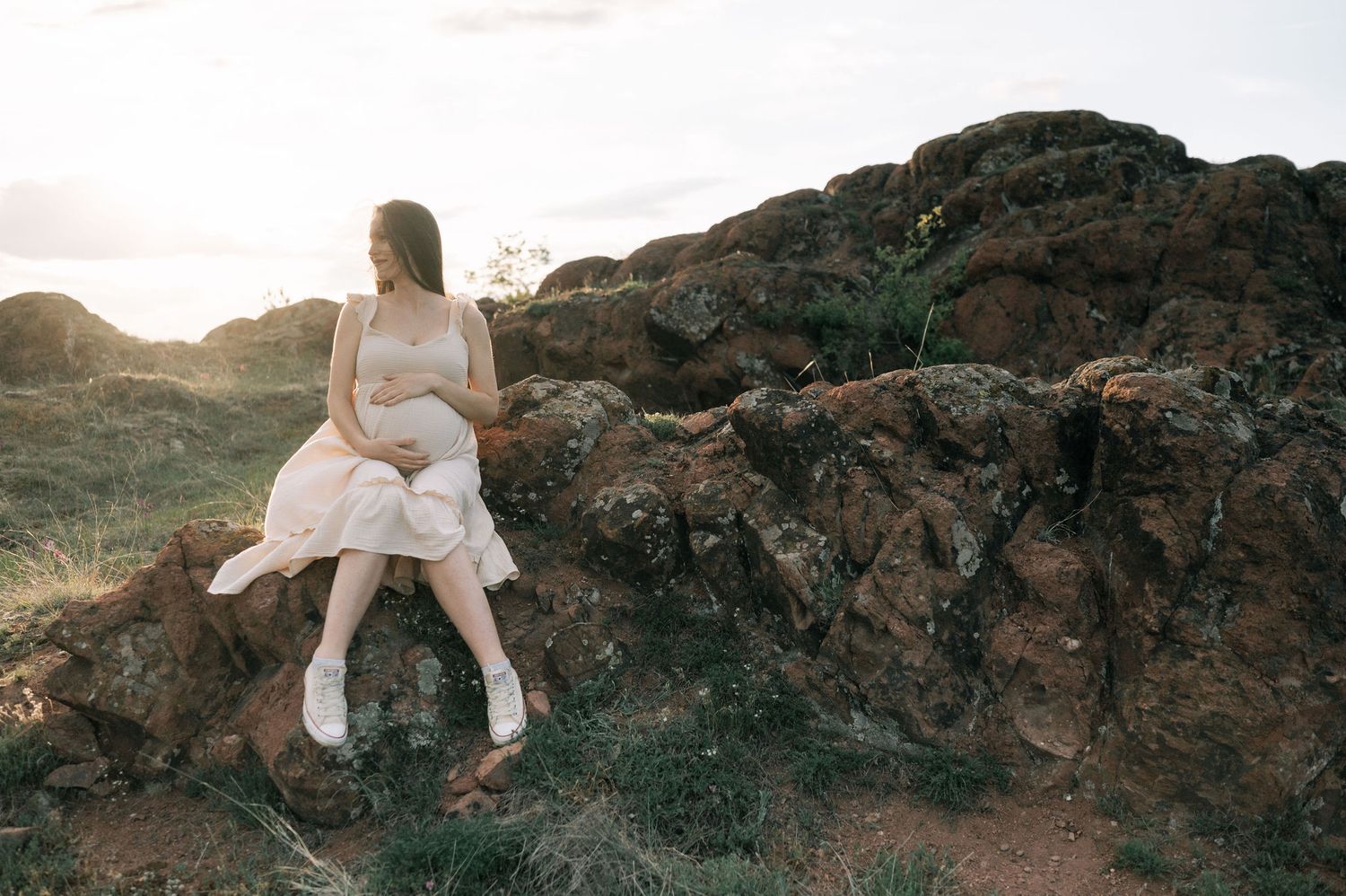 A person in a flowing white dress sits on dark rocky cliffs against a dramatic sunset backdrop.