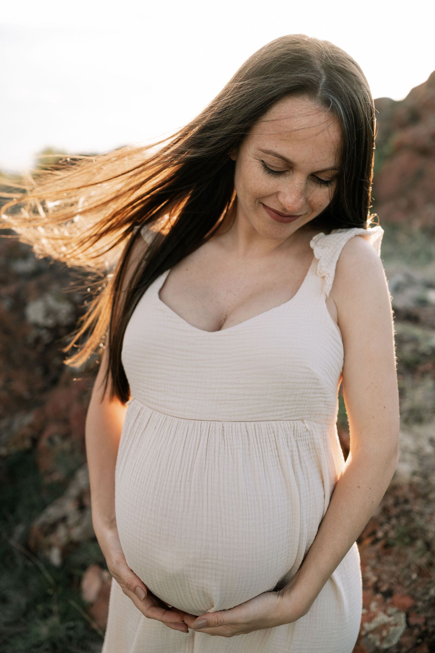 Woman in white dress poses outdoors with flowing hair in a series of maternity photos against a natural backdrop.