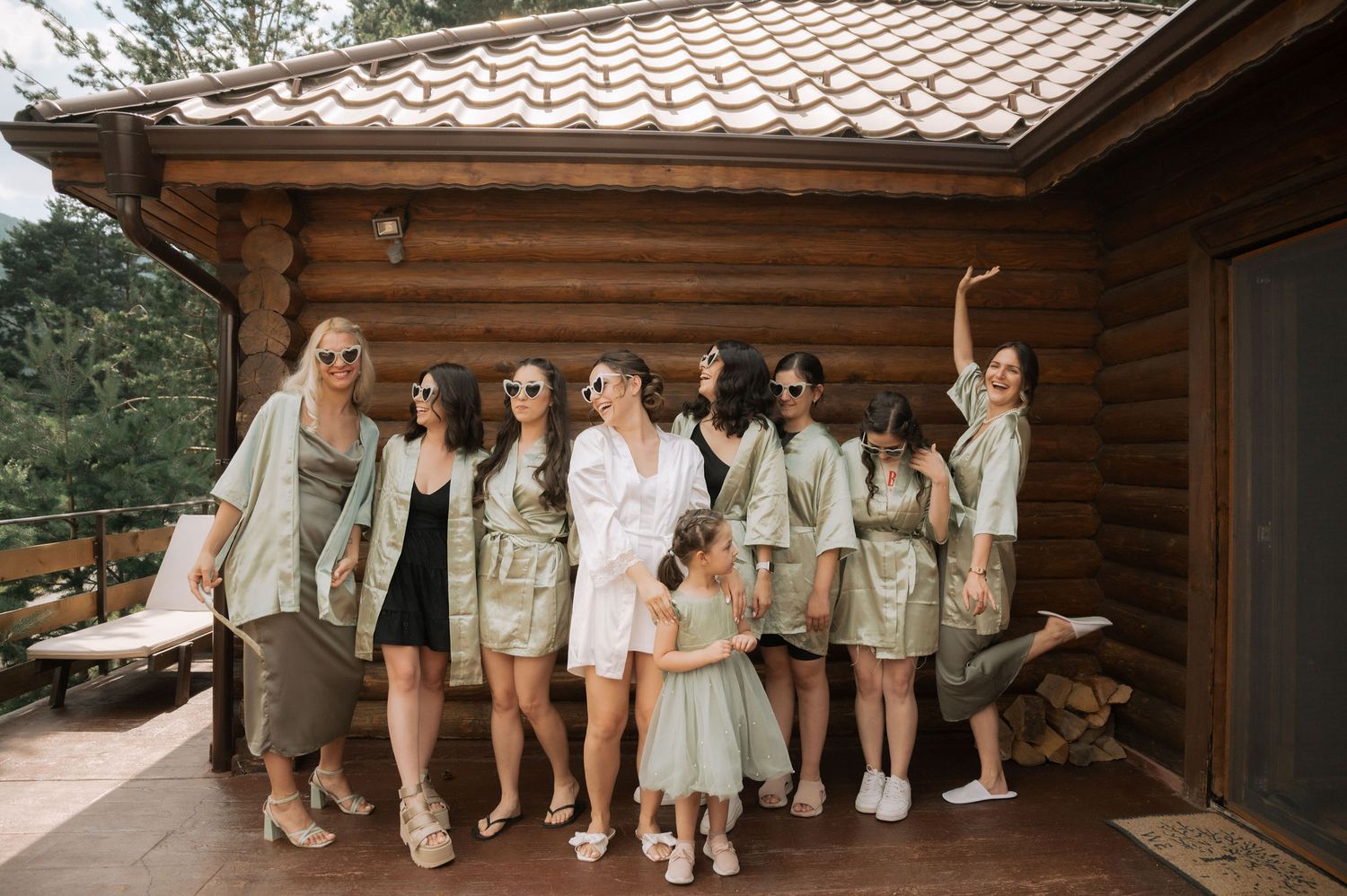 Wedding party in neutral colors poses together outside rustic log cabin venue.
