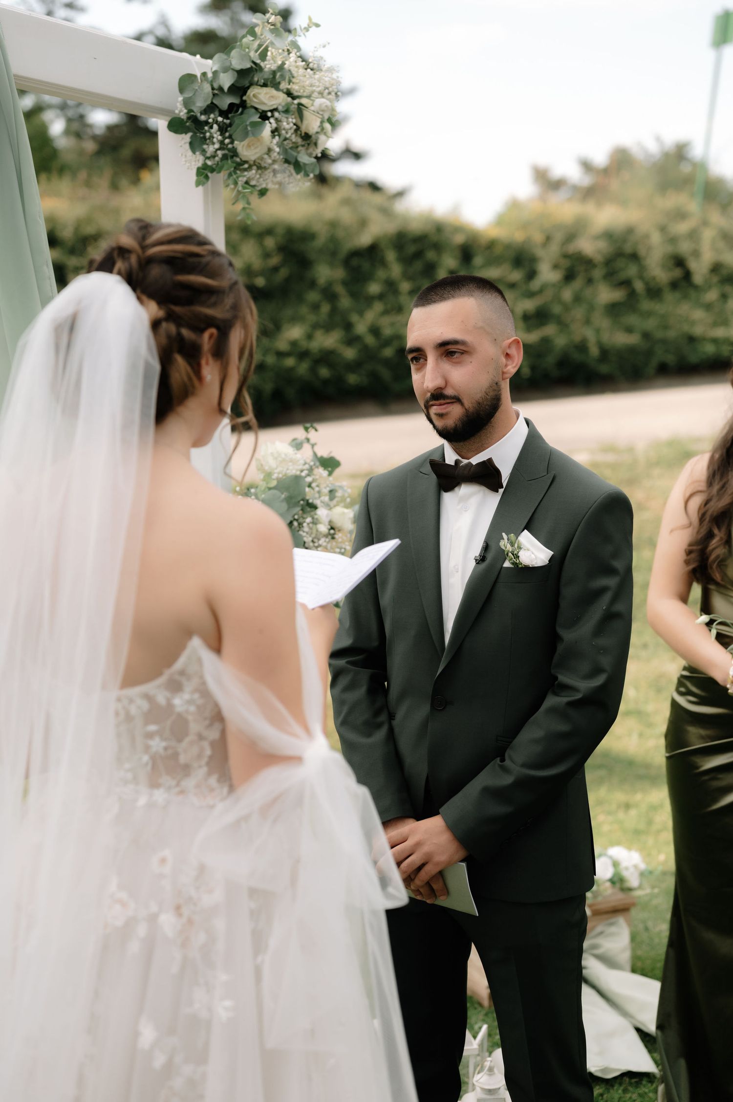 A couple exchanges vows during an outdoor wedding ceremony under a white draped gazebo with floral decorations.