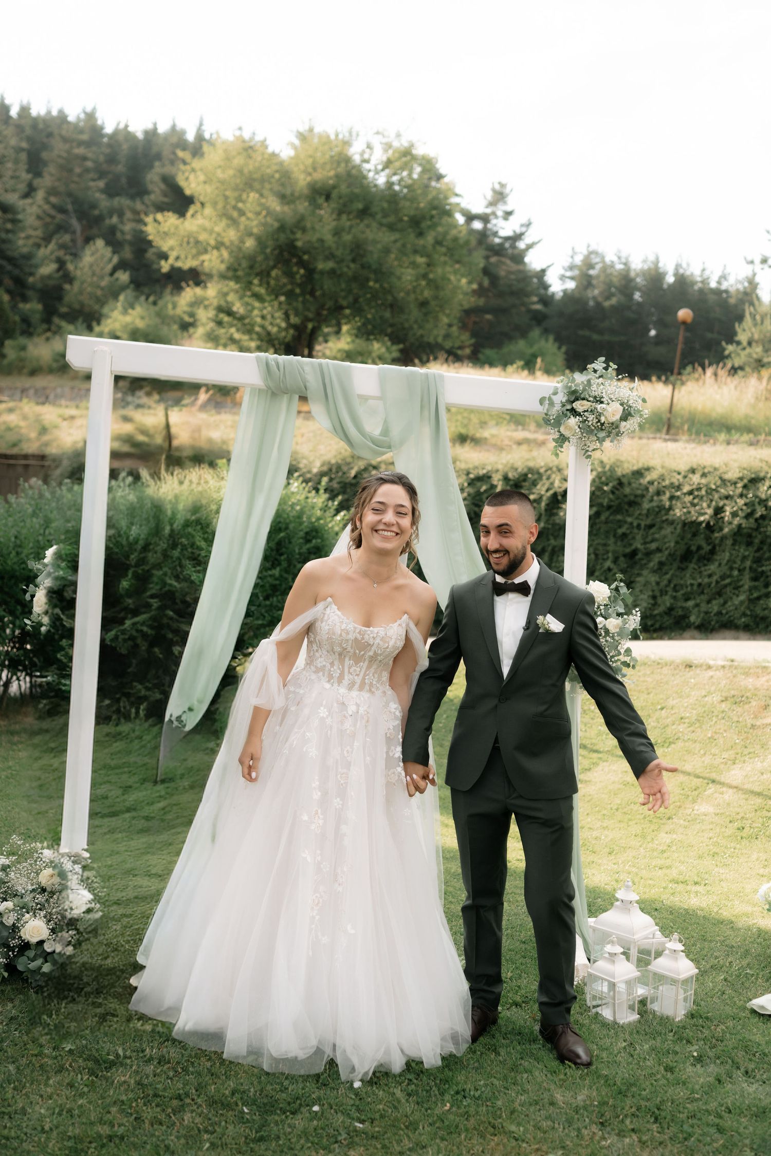 A wedding couple poses playfully in front of white draped curtains in an outdoor garden setting.
