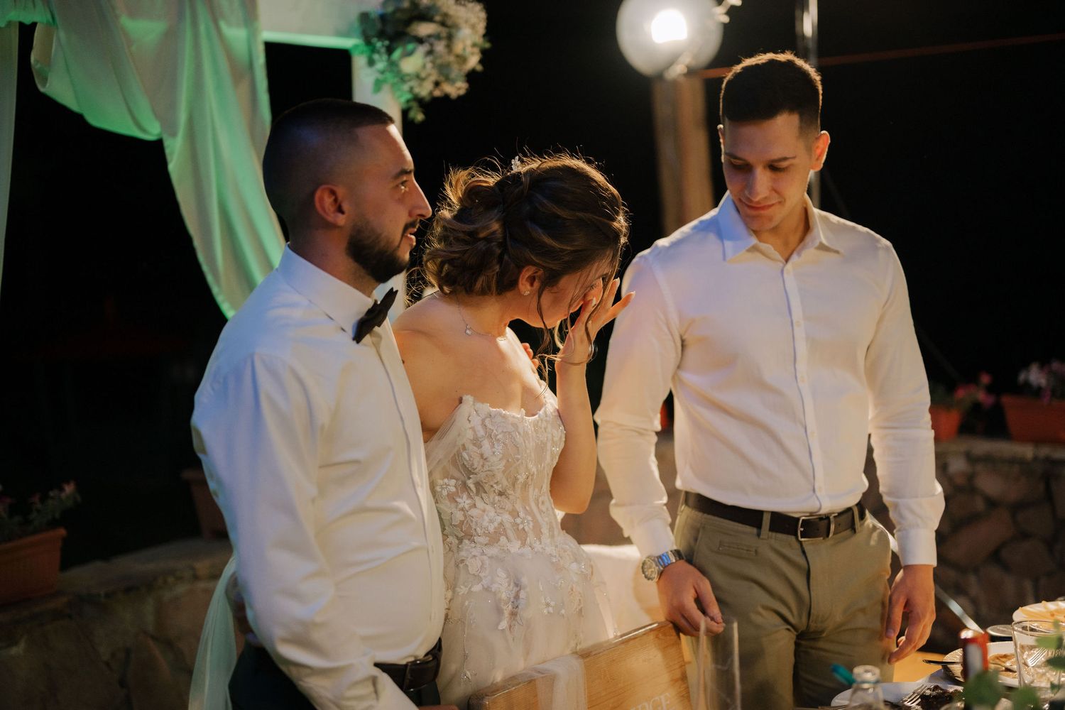 Three people in white attire share a candid moment during an evening wedding reception celebration.