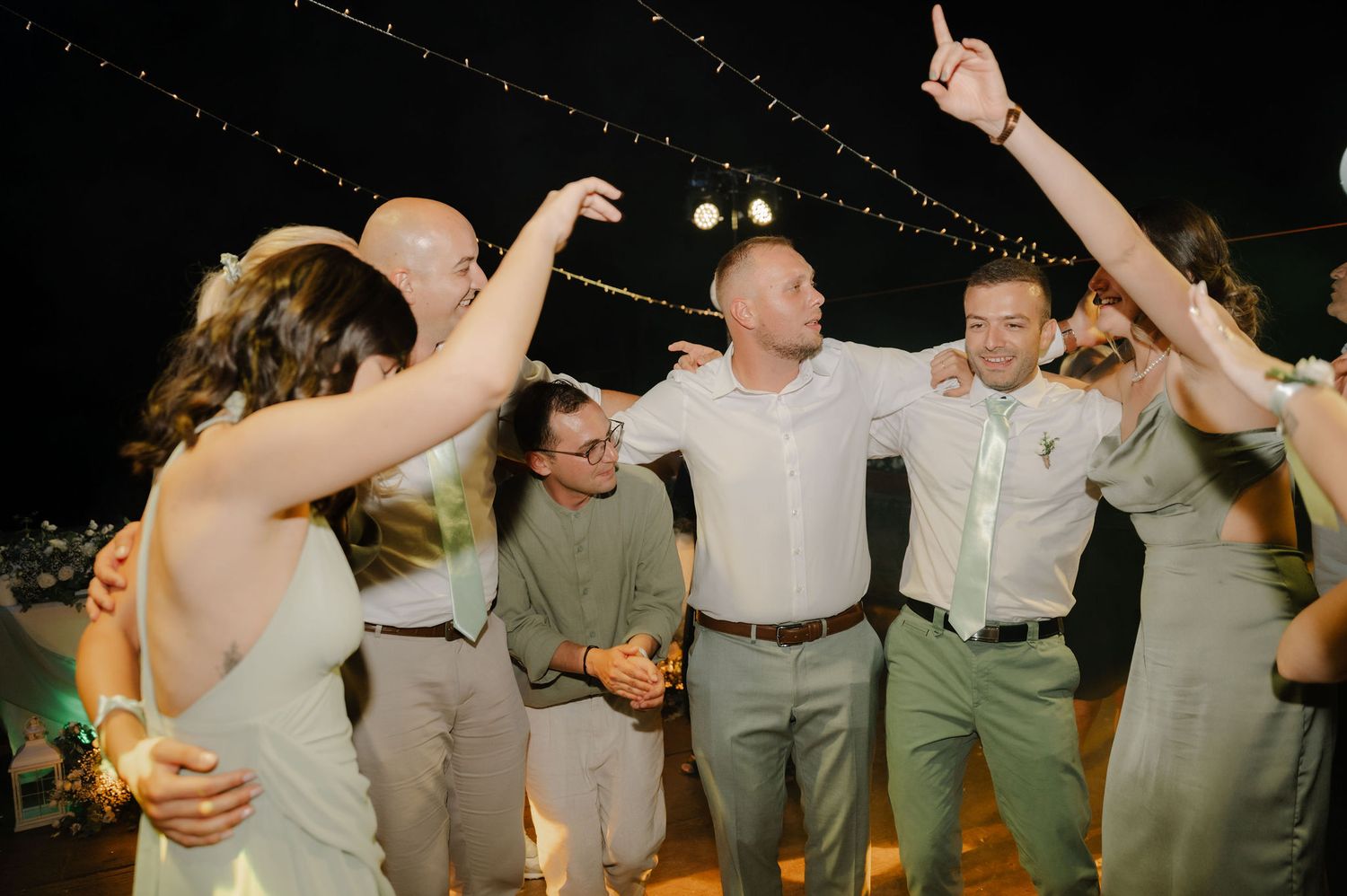 Wedding guests dance together in a line during a reception celebration with their arms around each other's shoulders.