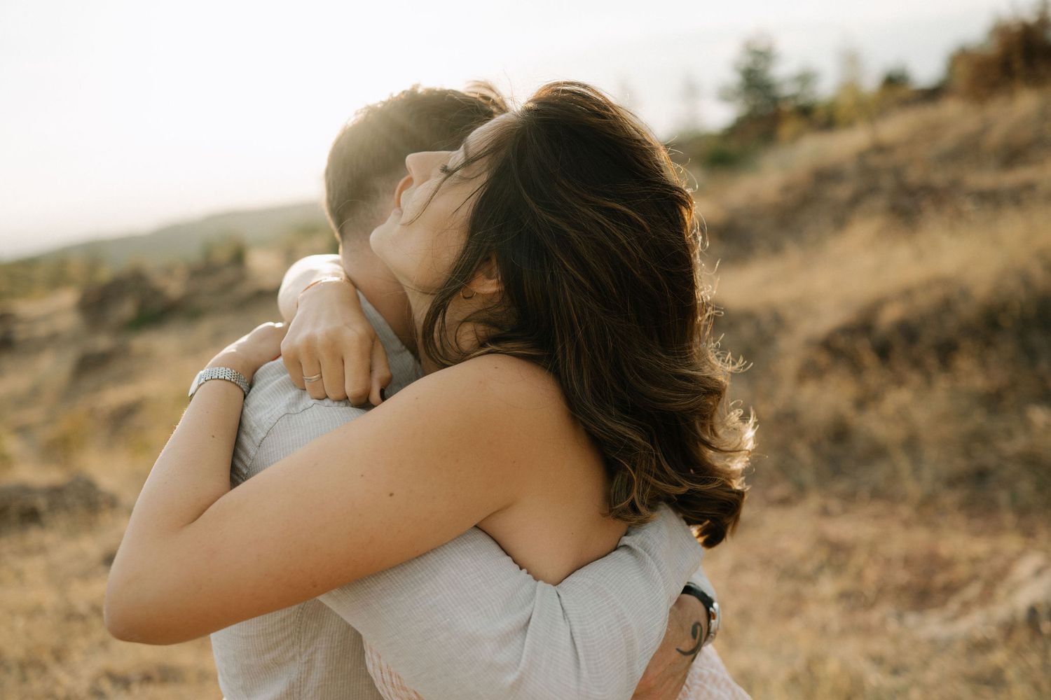 Two people embrace in a warm hug during golden hour on a hillside with soft sunlight illuminating the scene.