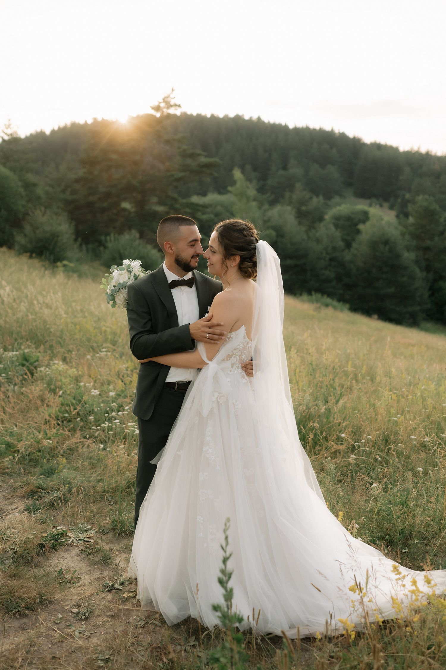 A romantic wedding couple embraces in a sunlit meadow during golden hour as her flowing white dress catches the light.