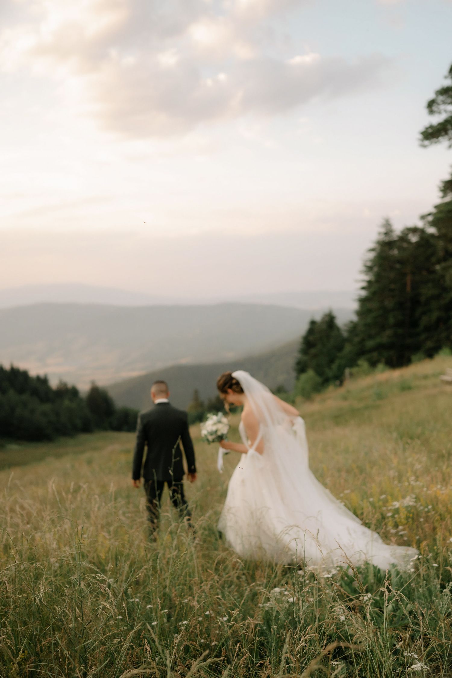 A couple in wedding attire walks through a scenic mountain meadow with tall grass at sunset.