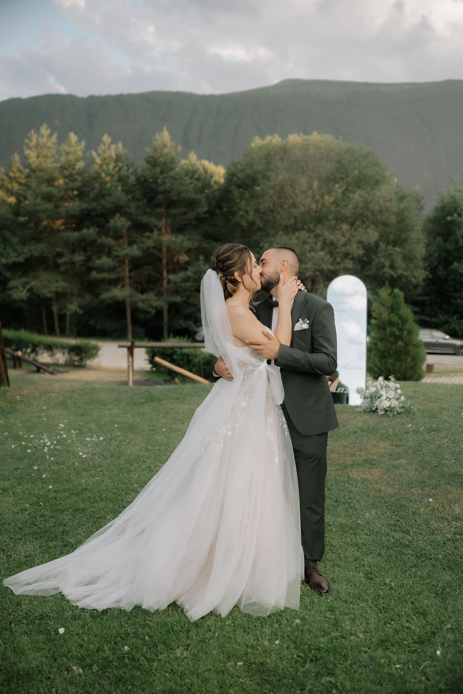 A romantic wedding couple shares a kiss on a grassy hillside with mountains and evergreen trees in the background.