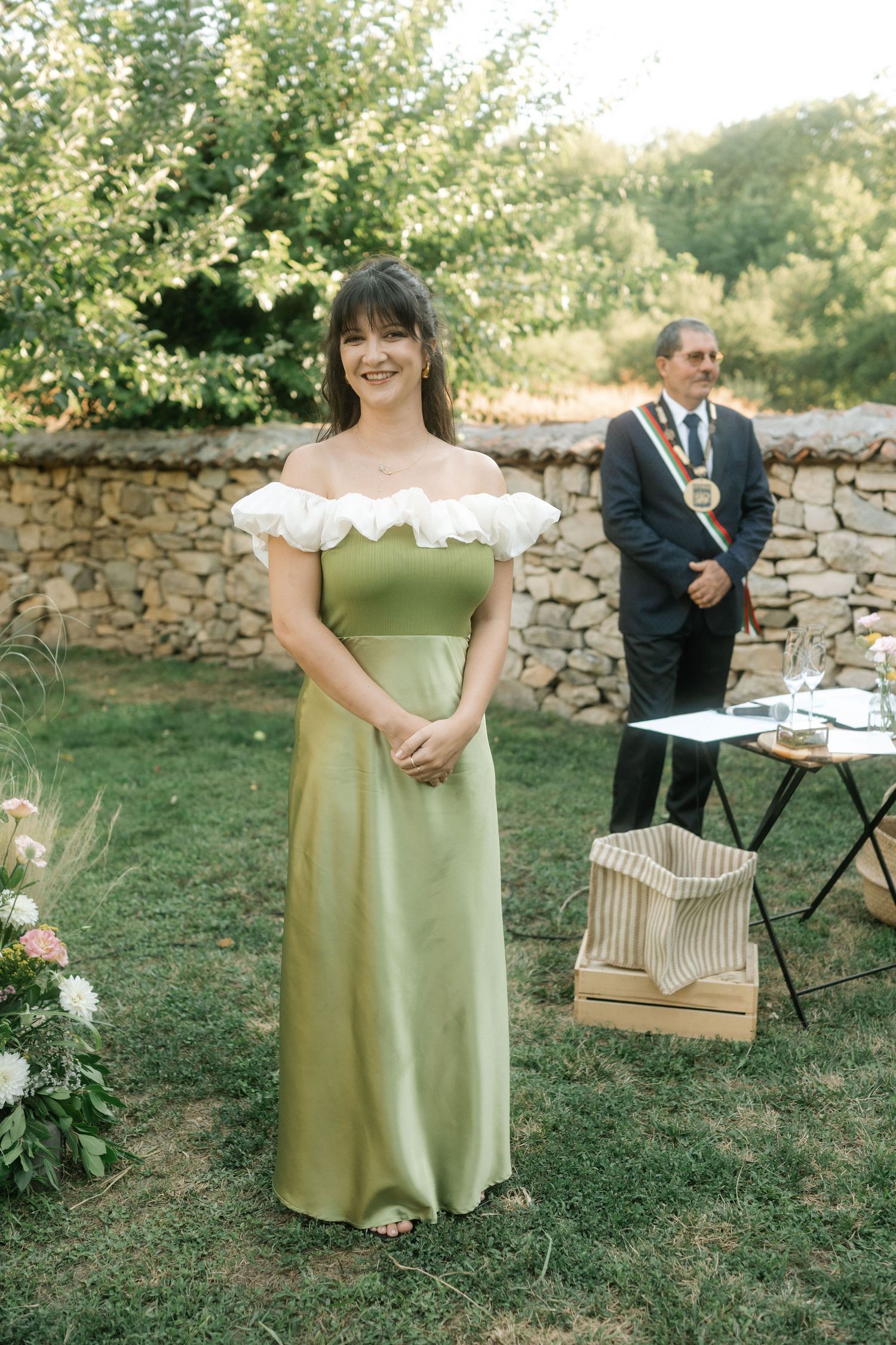 A person in a sage green dress stands in a garden setting near a rustic stone wall during an outdoor event.