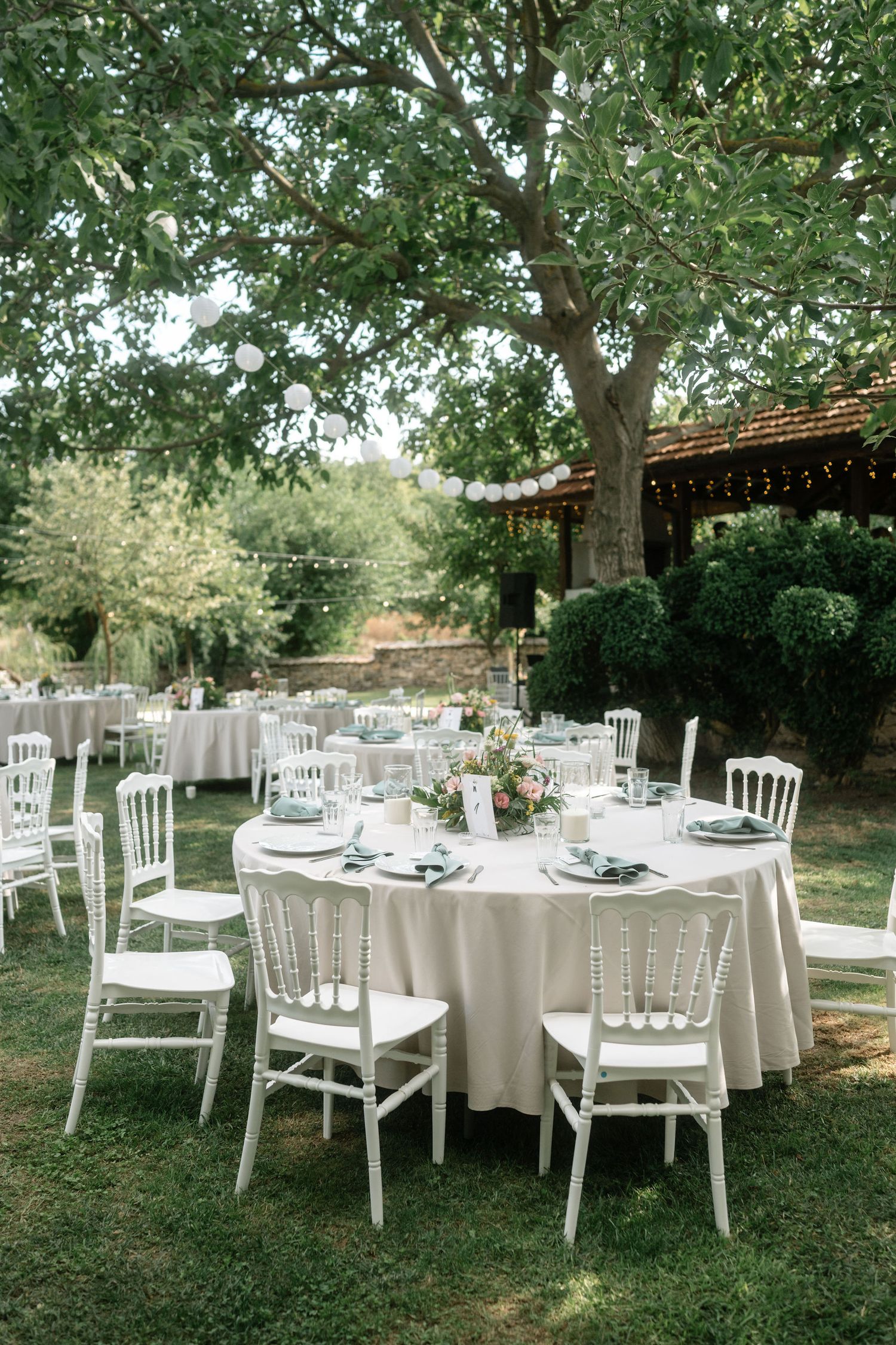 Elegant outdoor wedding reception setup with white round tables, chairs, and table settings under large shady trees.