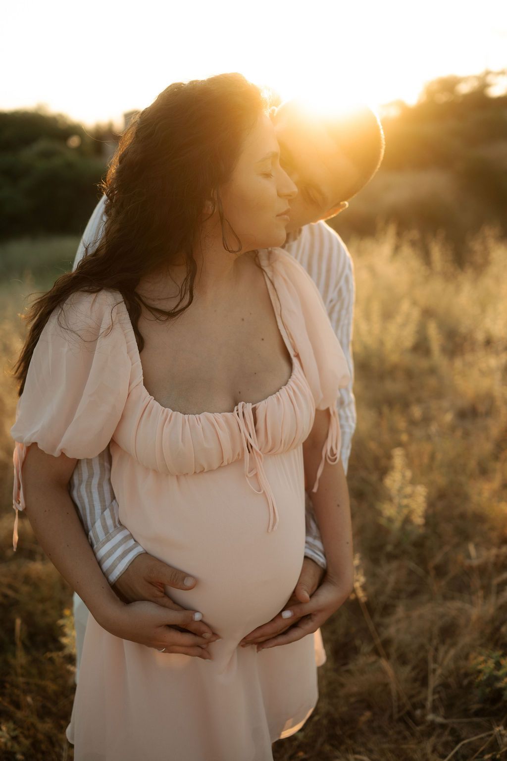 Maternity photoshoot in a golden field at sunset shows a flowing white dress and gentle hands cradling a baby bump.