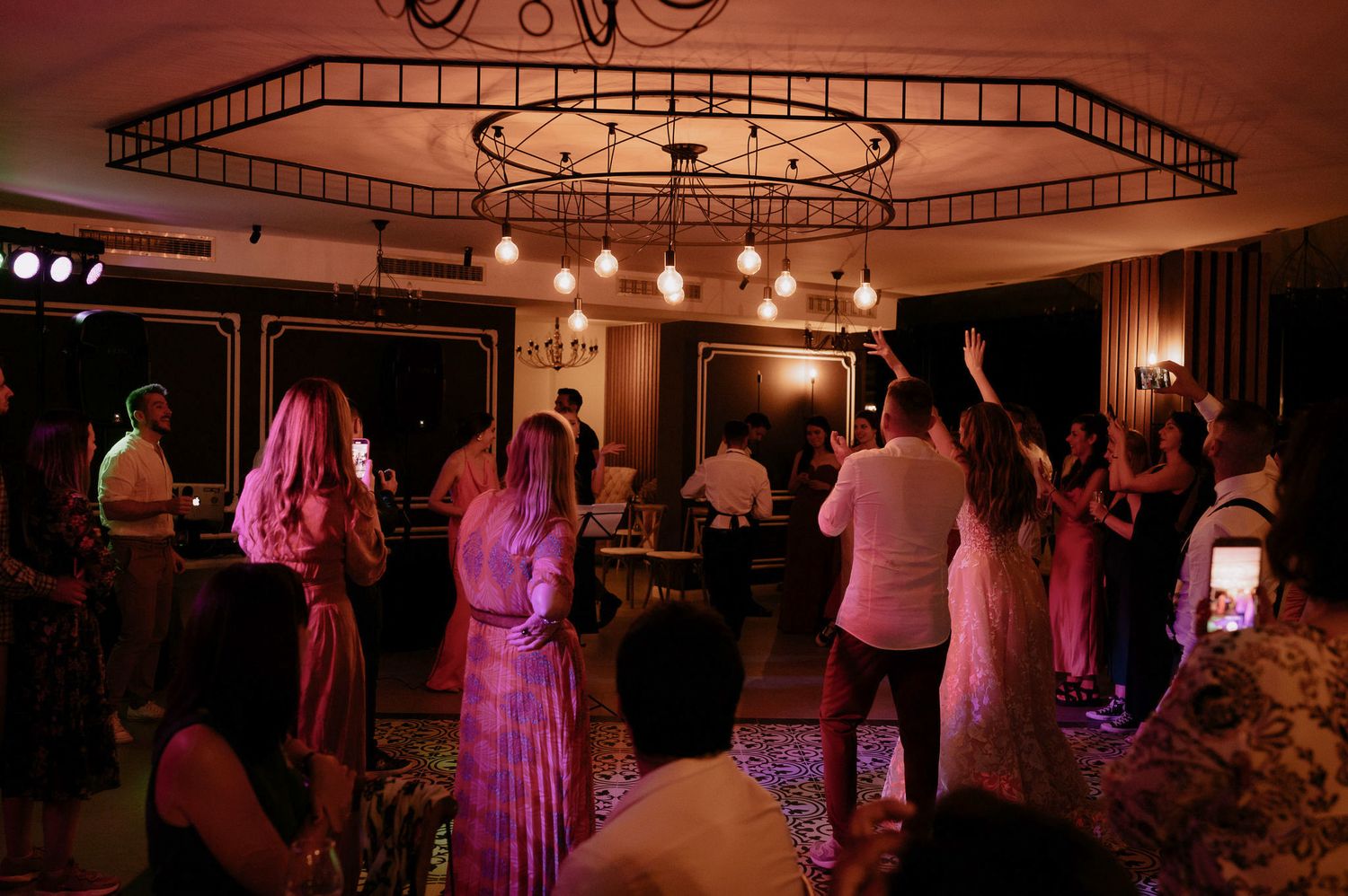 Wedding reception guests dancing on the dance floor under warm lighting and chandeliers at an elegant indoor venue.