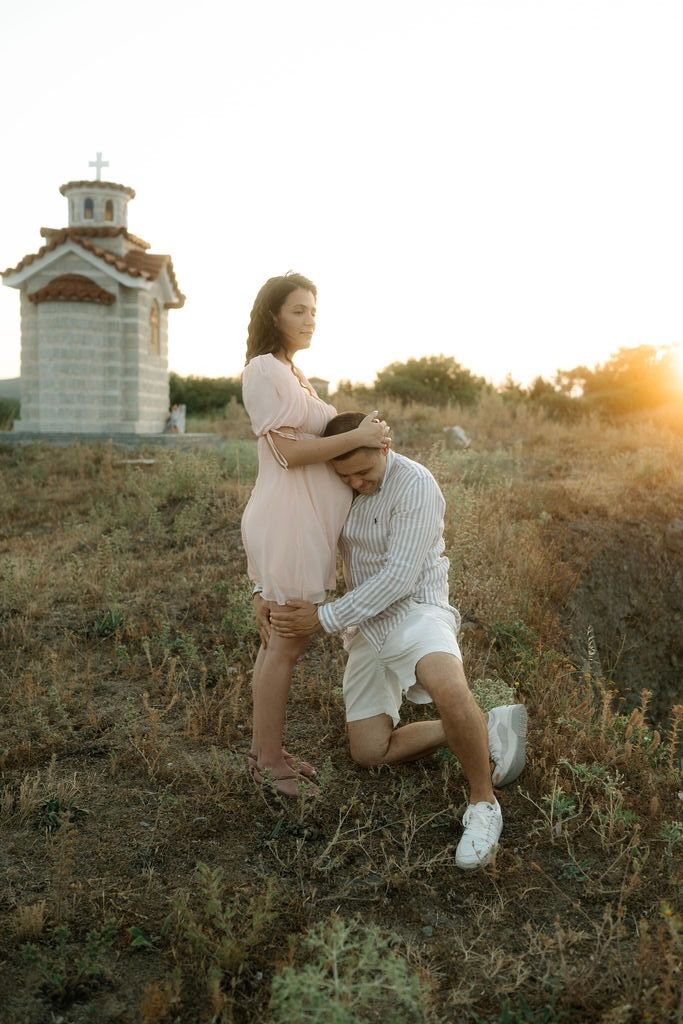 A couple in white outfits shares tender moments at sunset on a grassy hilltop with a church in the background.