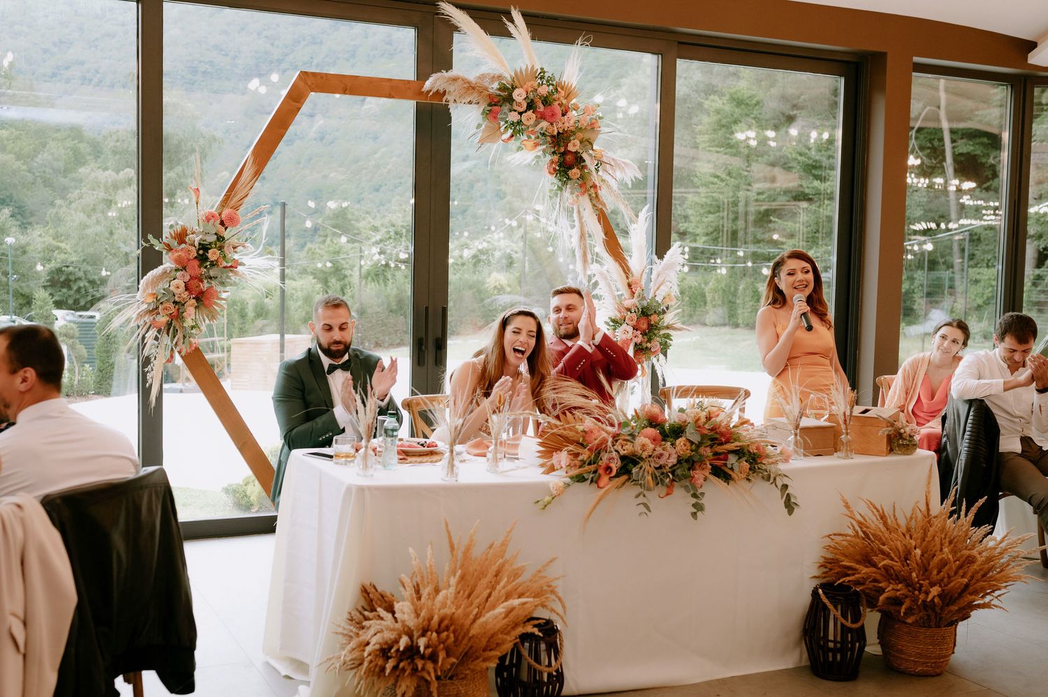 A wedding reception venue with glass walls showcasing natural scenery and elegant table settings decorated with dried flowers.