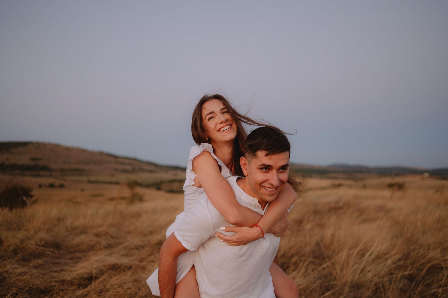 A couple shares a playful piggyback ride moment in a golden field at sunset.