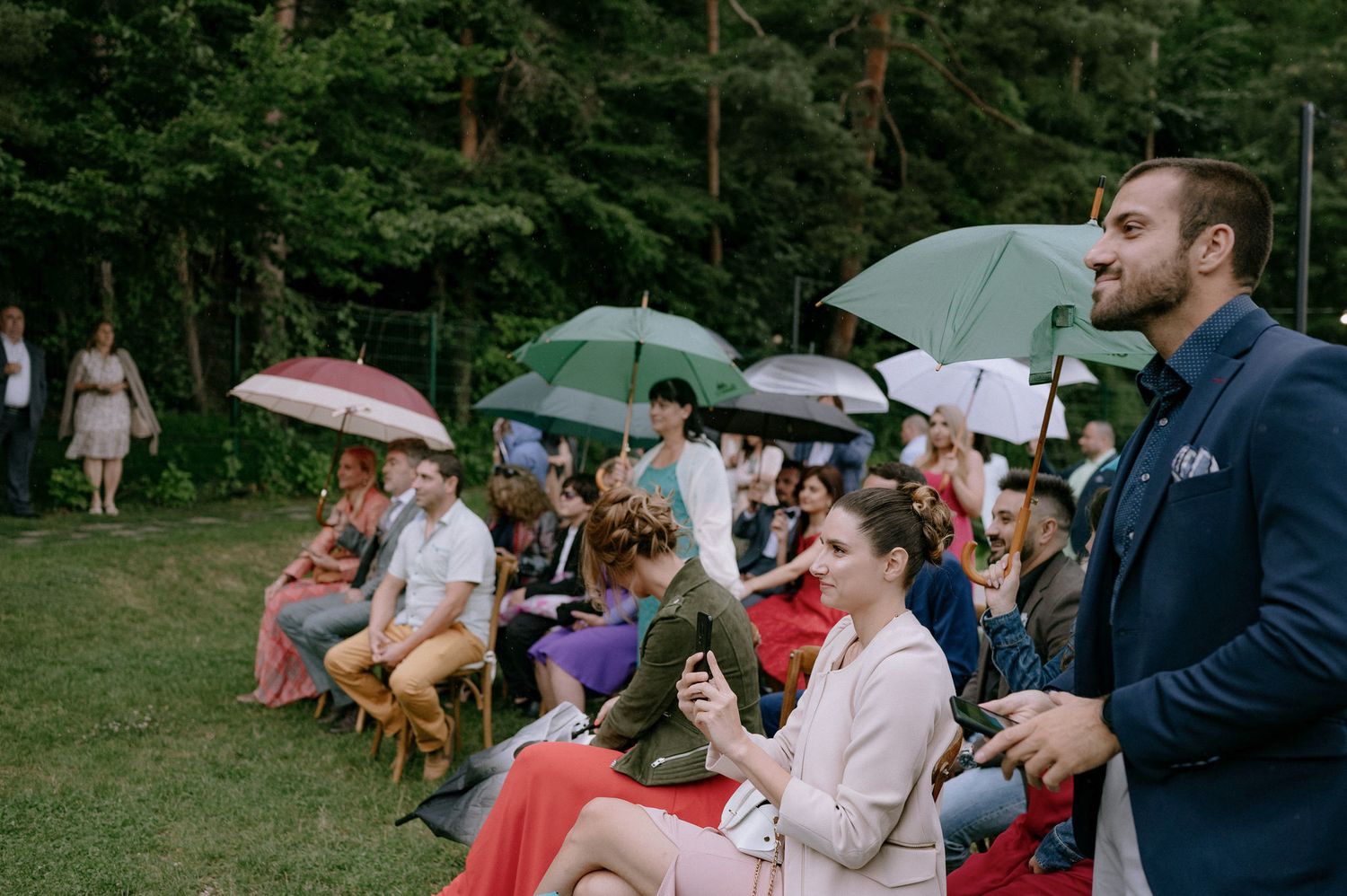 People sit on grass with umbrellas during an outdoor gathering in a park setting.