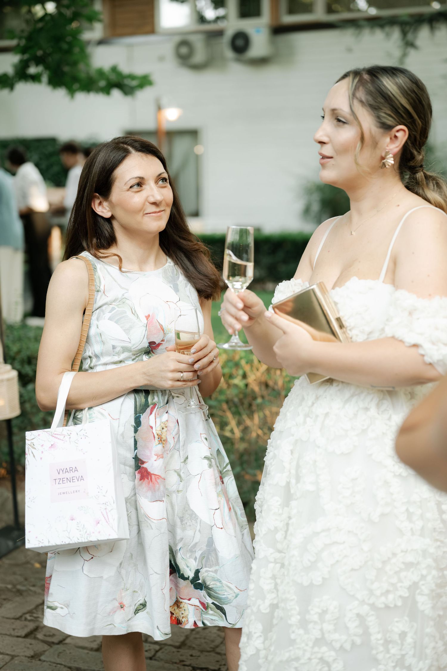 Two women in white dresses share a joyful moment while holding champagne glasses and gift bags at an outdoor event.