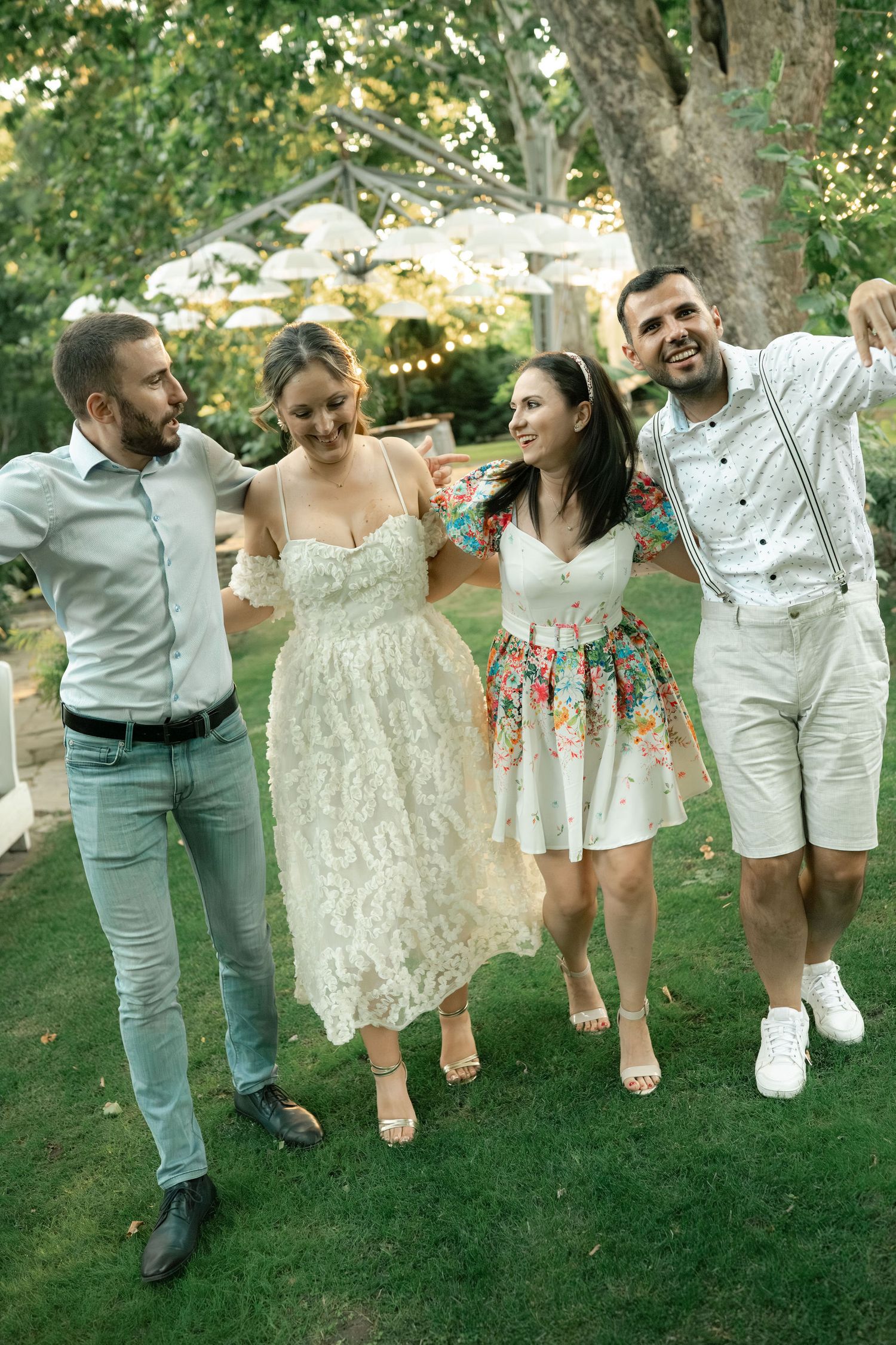A group of friends in white and light-colored outfits walking and laughing together on a grassy lawn in summer.