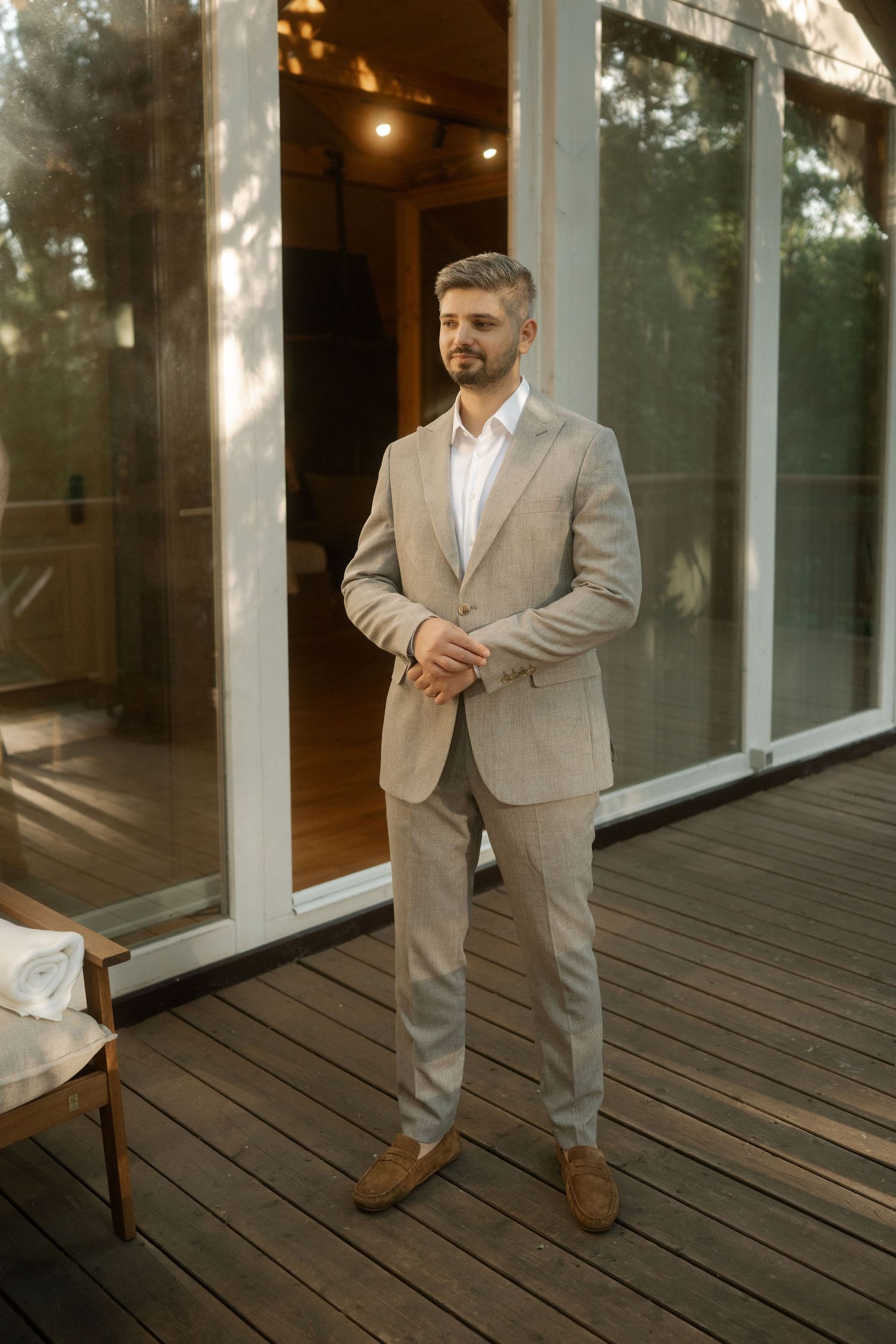 Professional wearing a beige suit stands on a wooden deck outside glass windows in warm evening lighting.
