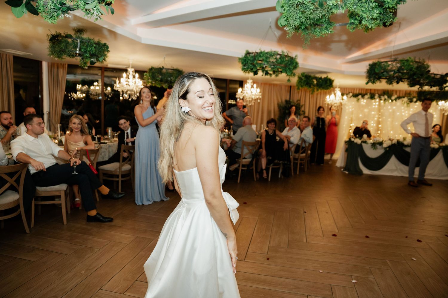 Elegant wedding reception hall decorated with greenery and chandeliers as guests dance and celebrate on wooden floors.