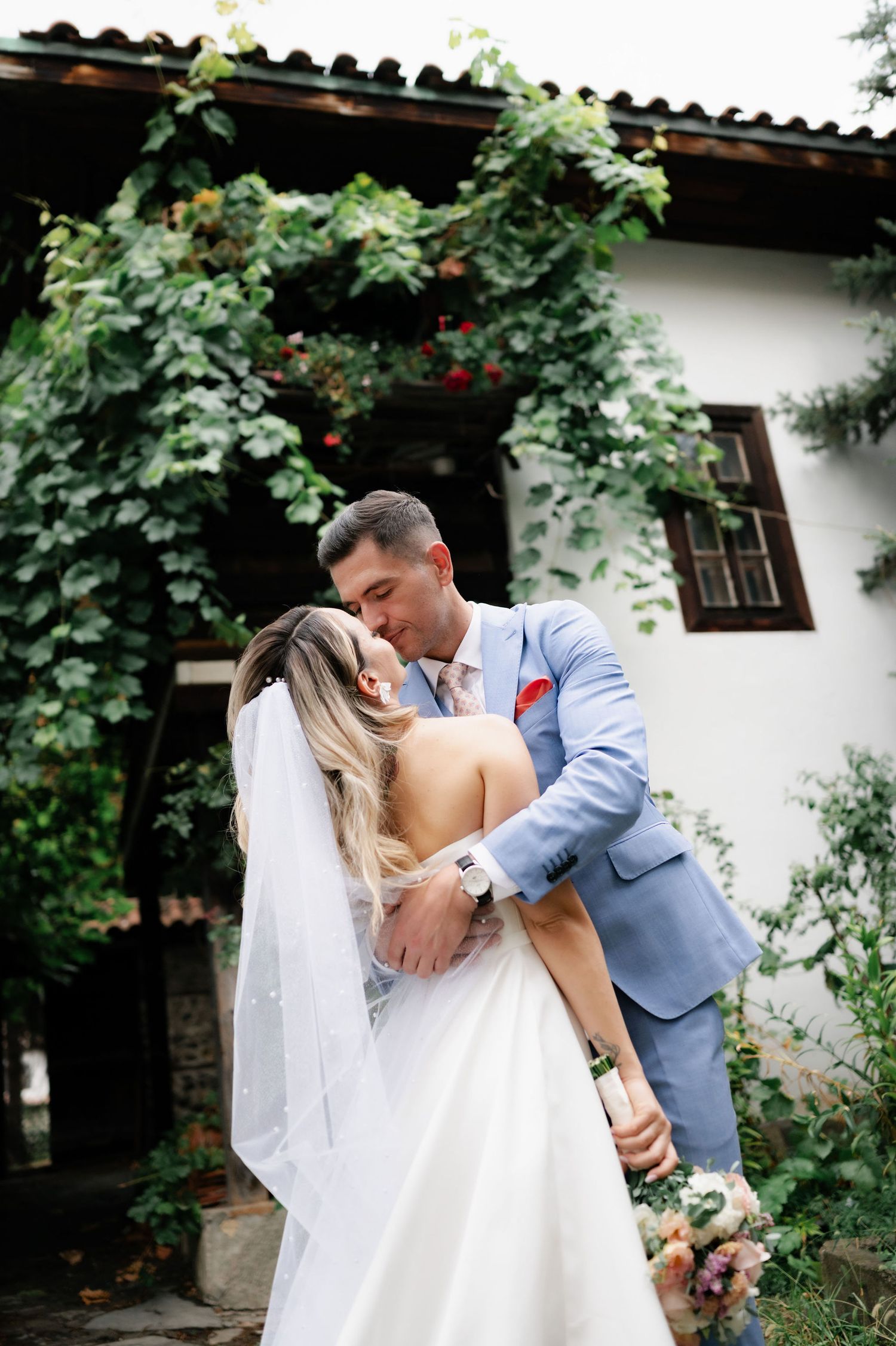 Romantic newlywed couple shares intimate moments in a garden setting with white walls and green foliage.