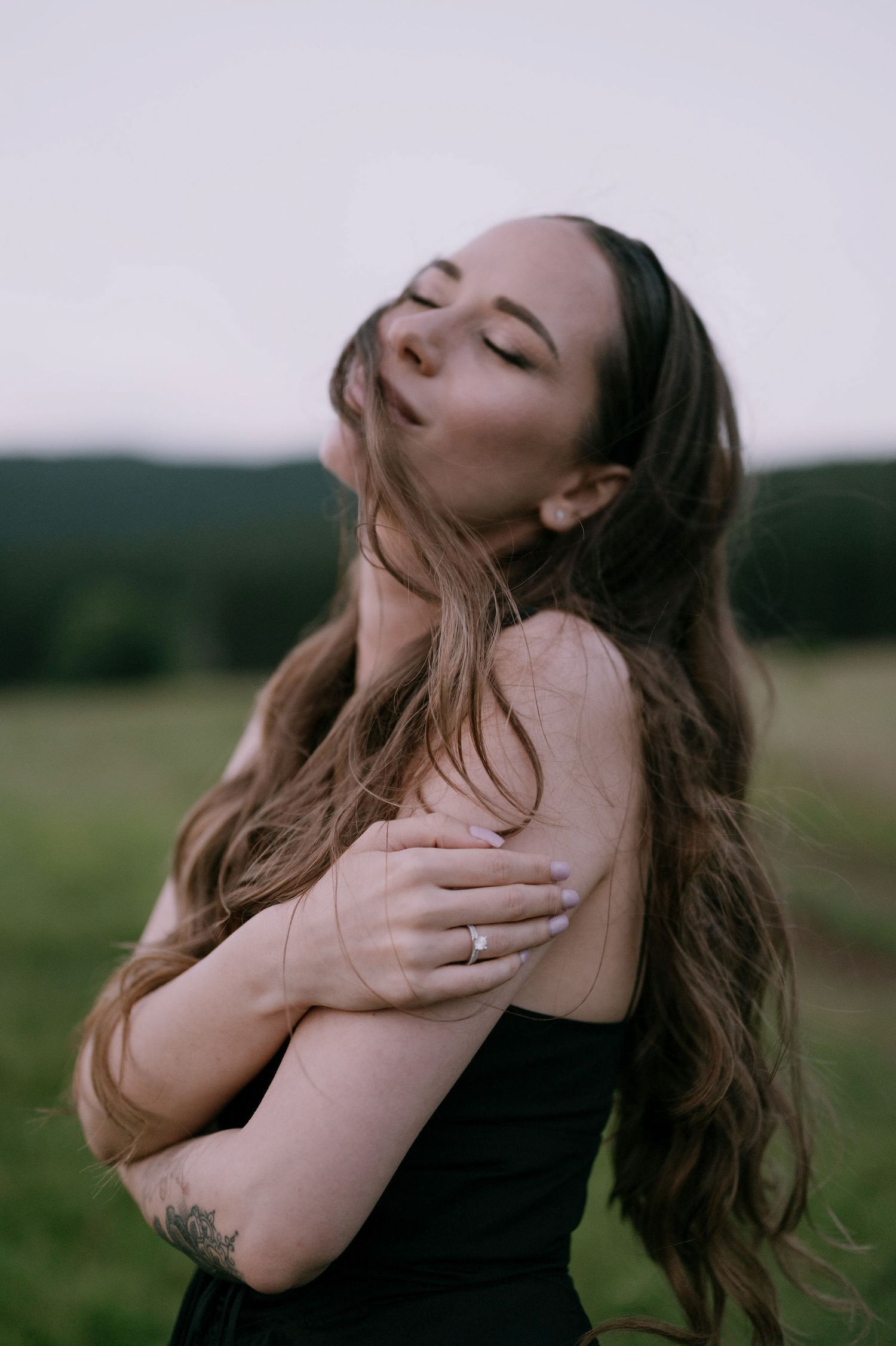 A person with long brown hair stands in a field with eyes closed, wearing a black top in moody outdoor lighting.