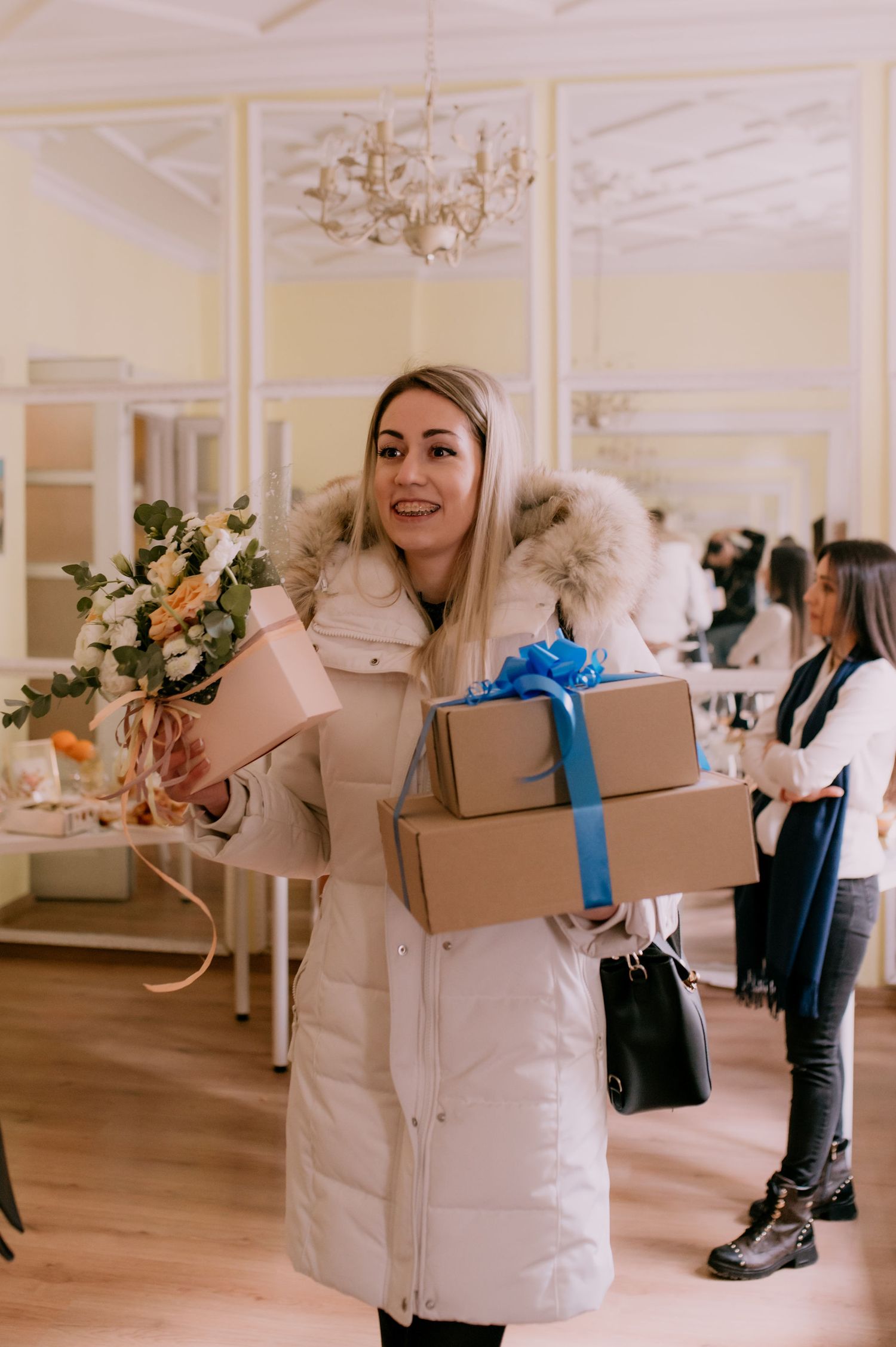 Person in a white winter coat holding gift boxes and flowers in an elegant room with a chandelier.