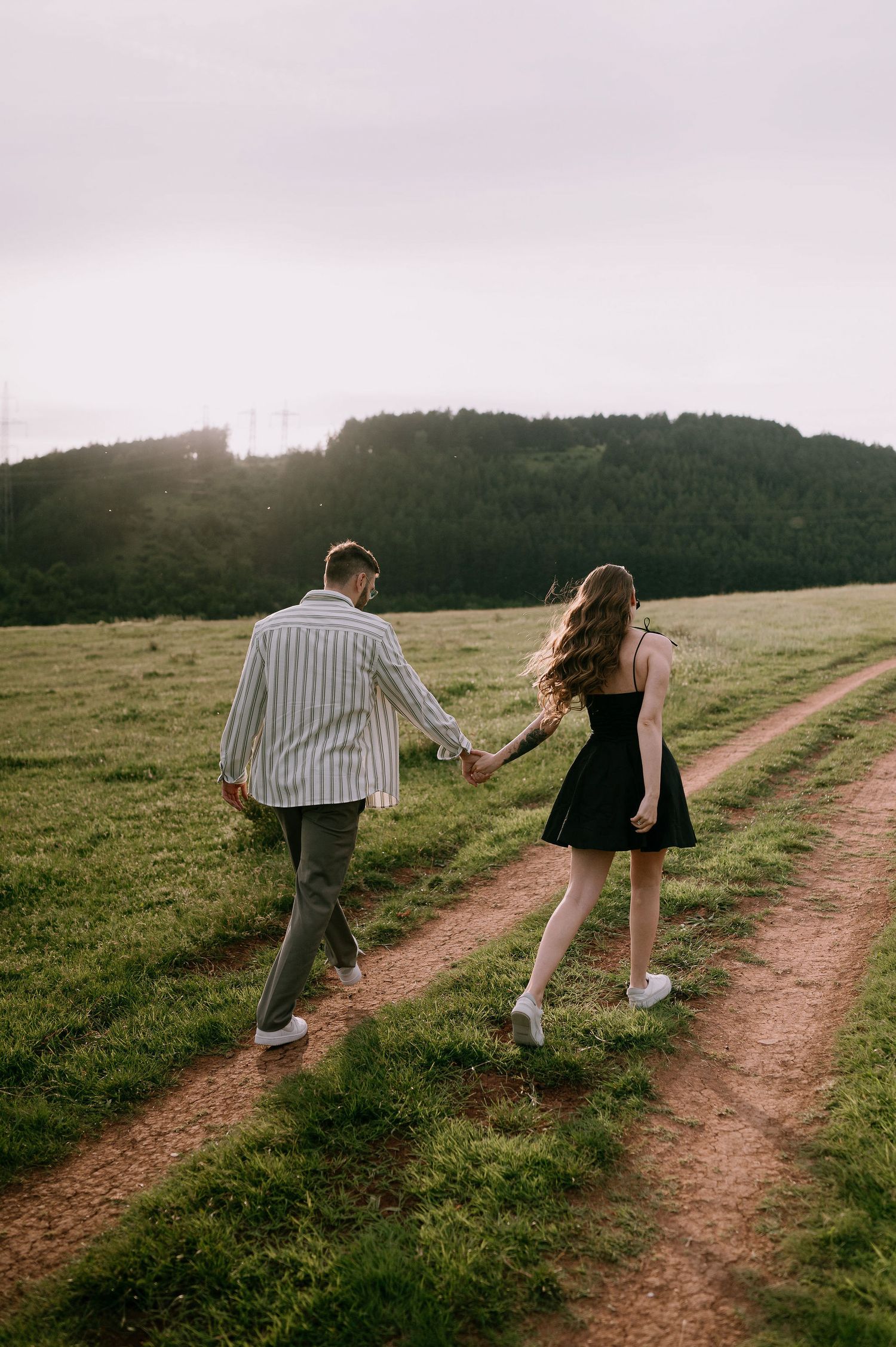 Two people walk hand in hand along a scenic dirt path through a grassy hillside at sunset.