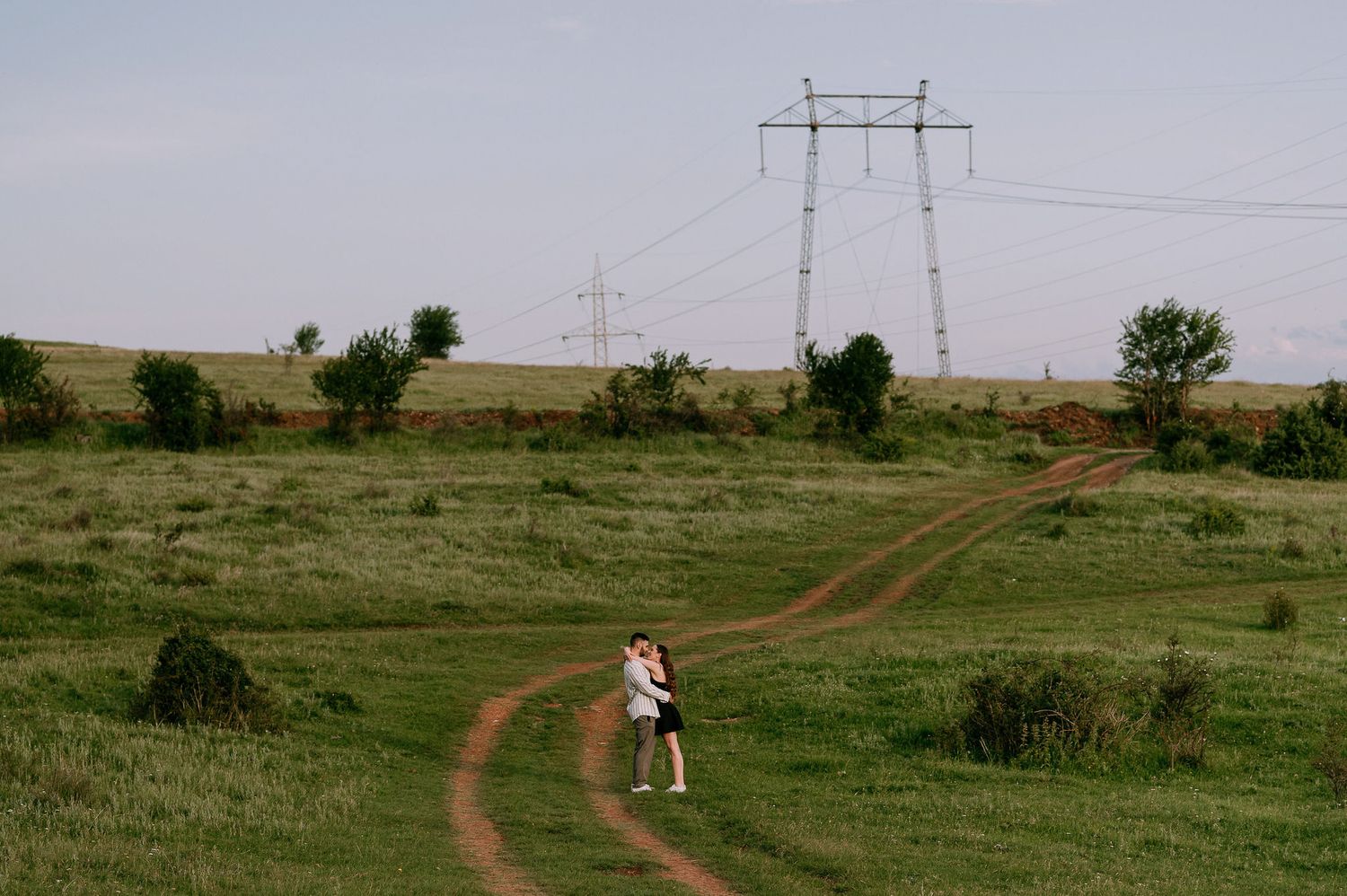 A winding dirt path leads through green grassland with electrical transmission towers stretching into the distance.