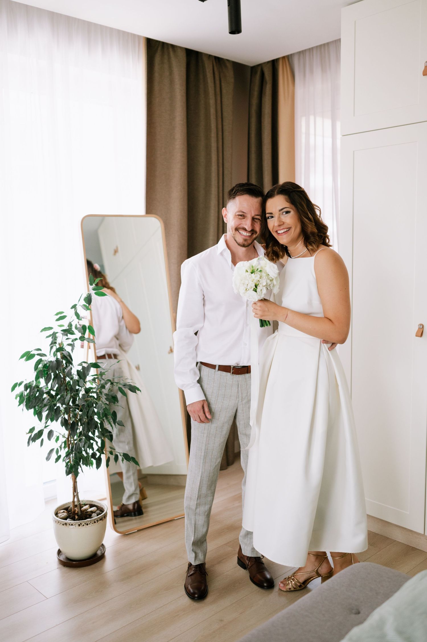 A couple shares joyful moments in a bright minimalist room with white walls and a potted plant.