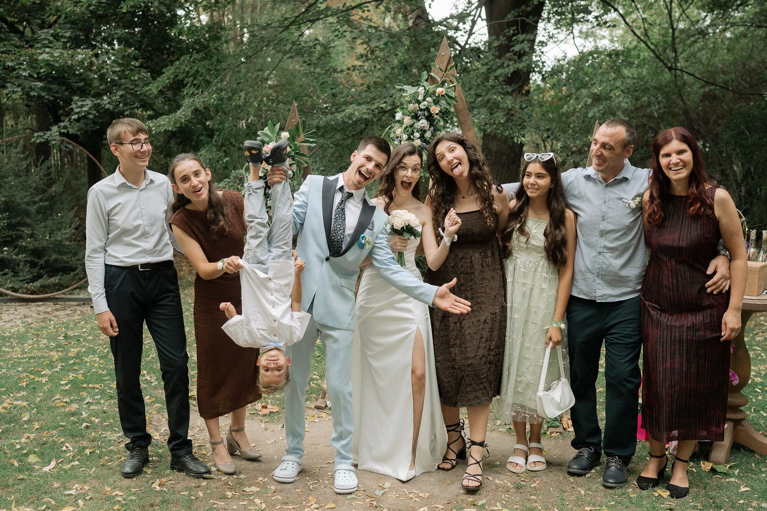 Group of wedding guests pose together outdoors in coordinated brown and white outfits in a garden setting.