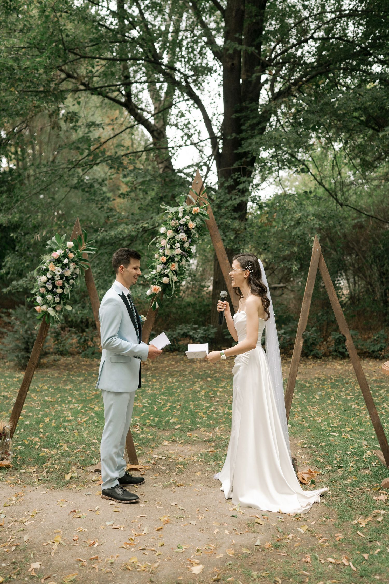 A couple shares a playful moment on wooden swings in a wooded outdoor setting surrounded by fallen autumn leaves.