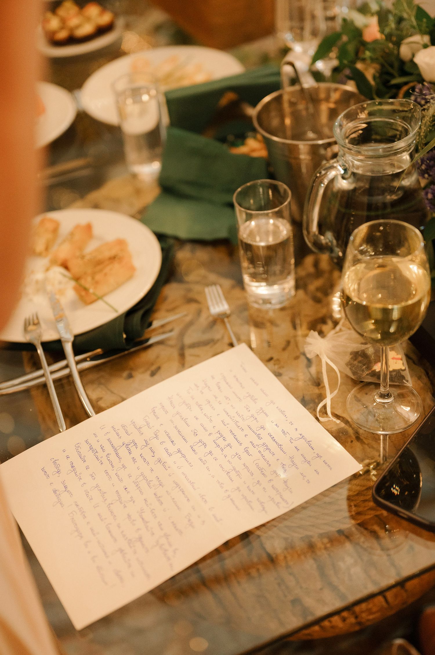 A handwritten note sits on a rustic wooden table alongside wine glasses and dinner plates in warm lighting.