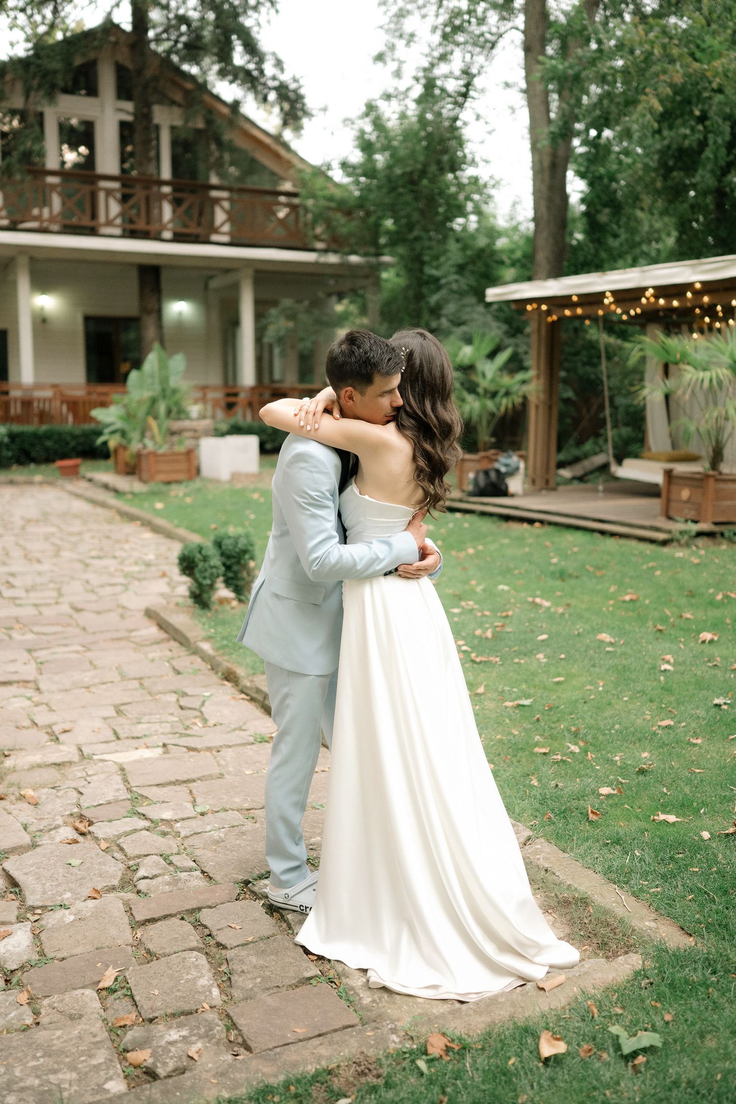 A couple in wedding attire dips and dances together on a stone patio surrounded by lush greenery.