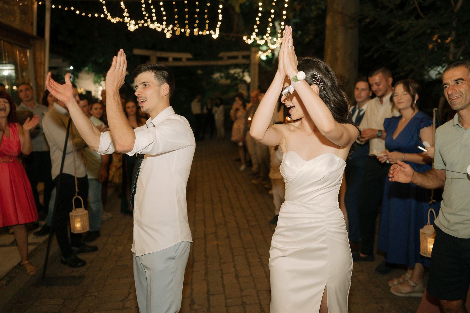 Wedding guests dance and celebrate on a rustic outdoor patio decorated with string lights above.