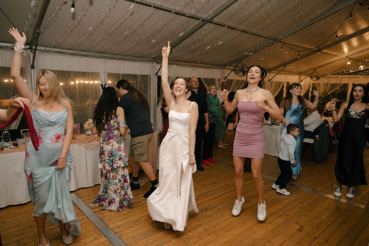 People dancing and celebrating at a lively wedding reception in a rustic indoor venue with wooden ceiling beams.