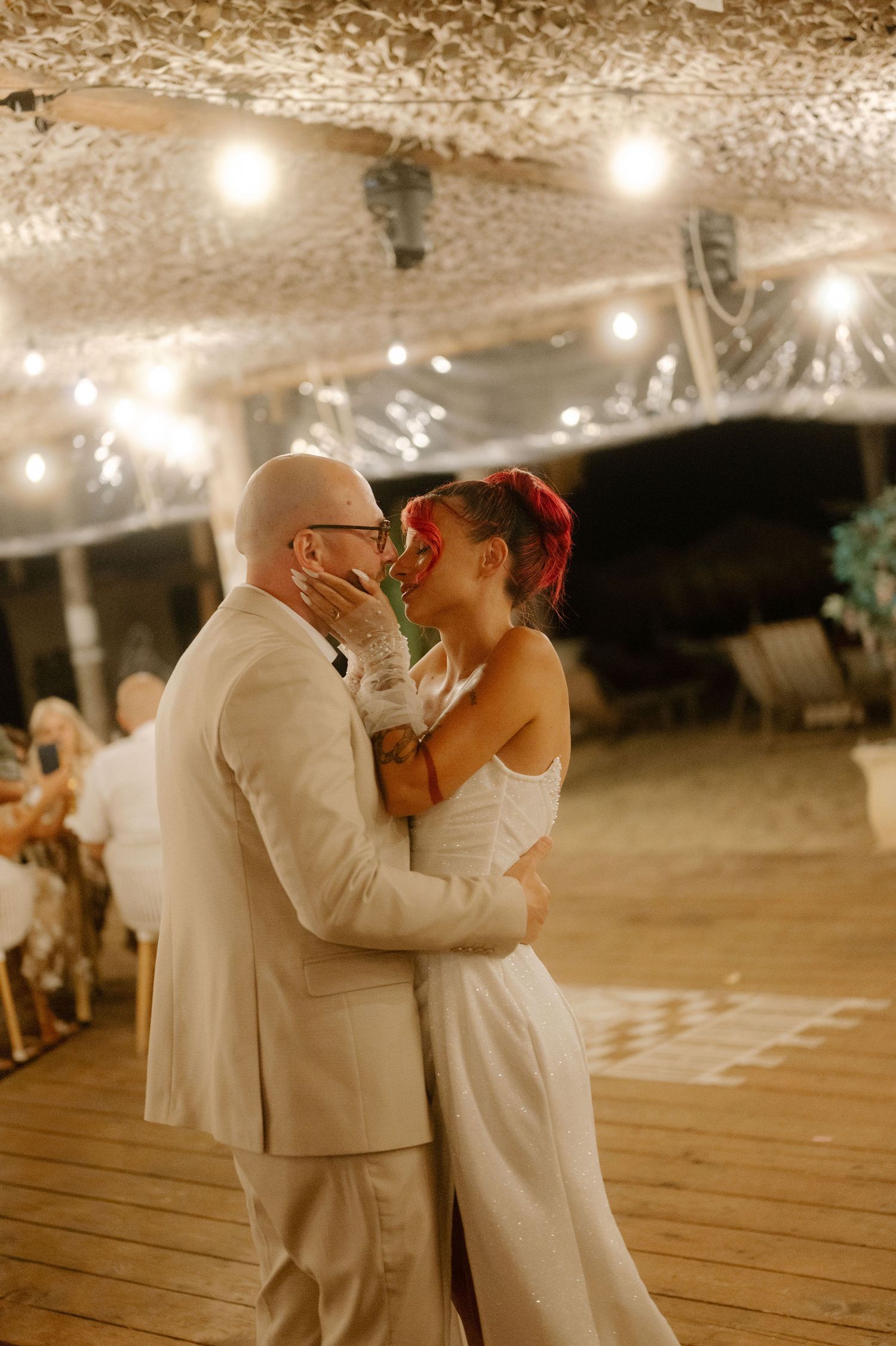 A couple shares their first dance as newlyweds under twinkling string lights on a wooden deck at dusk.