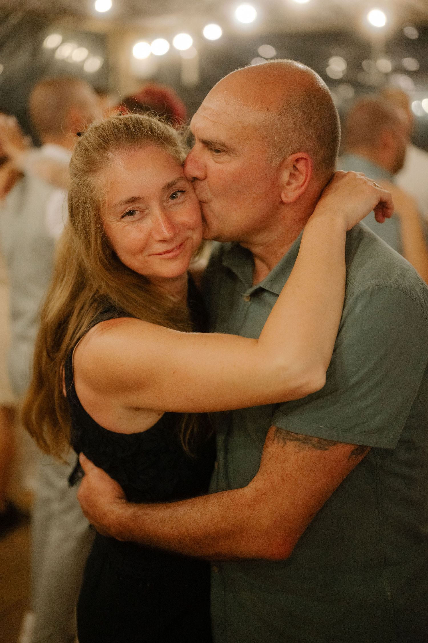 Two people share an affectionate embrace while dancing at an indoor event with warm ambient lighting.