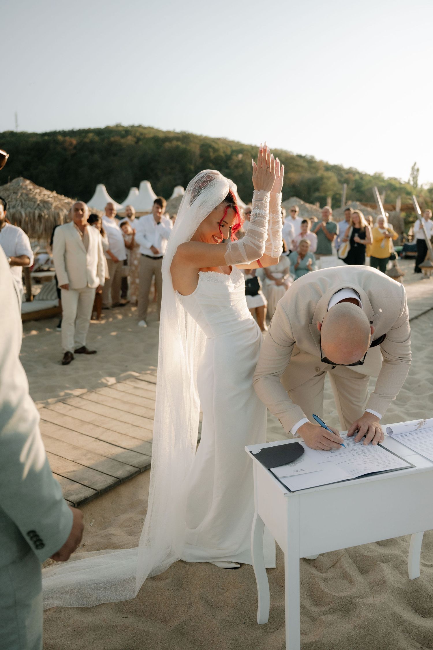 Wedding guests in white attire sit on beach chairs arranged in rows during an outdoor beach ceremony at sunset.