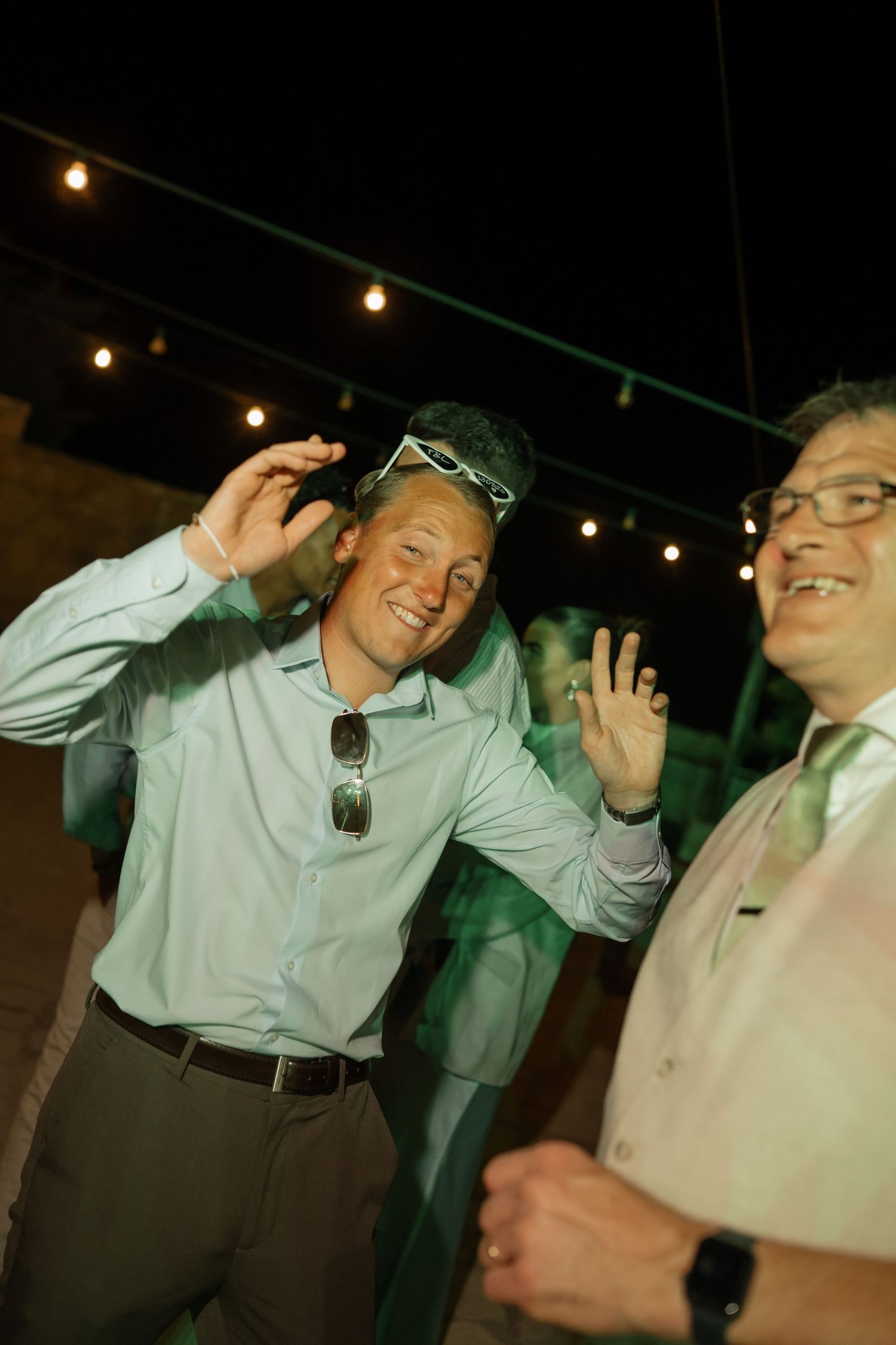 Two men dancing and smiling at an outdoor evening party with string lights overhead.